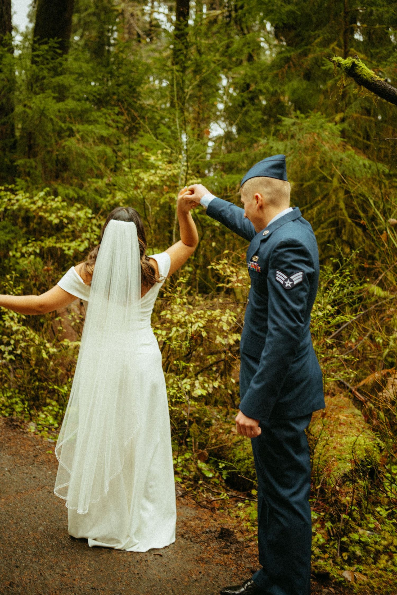 A military service member and bride share a romantic dance moment on a wooded path beneath evergreen trees.