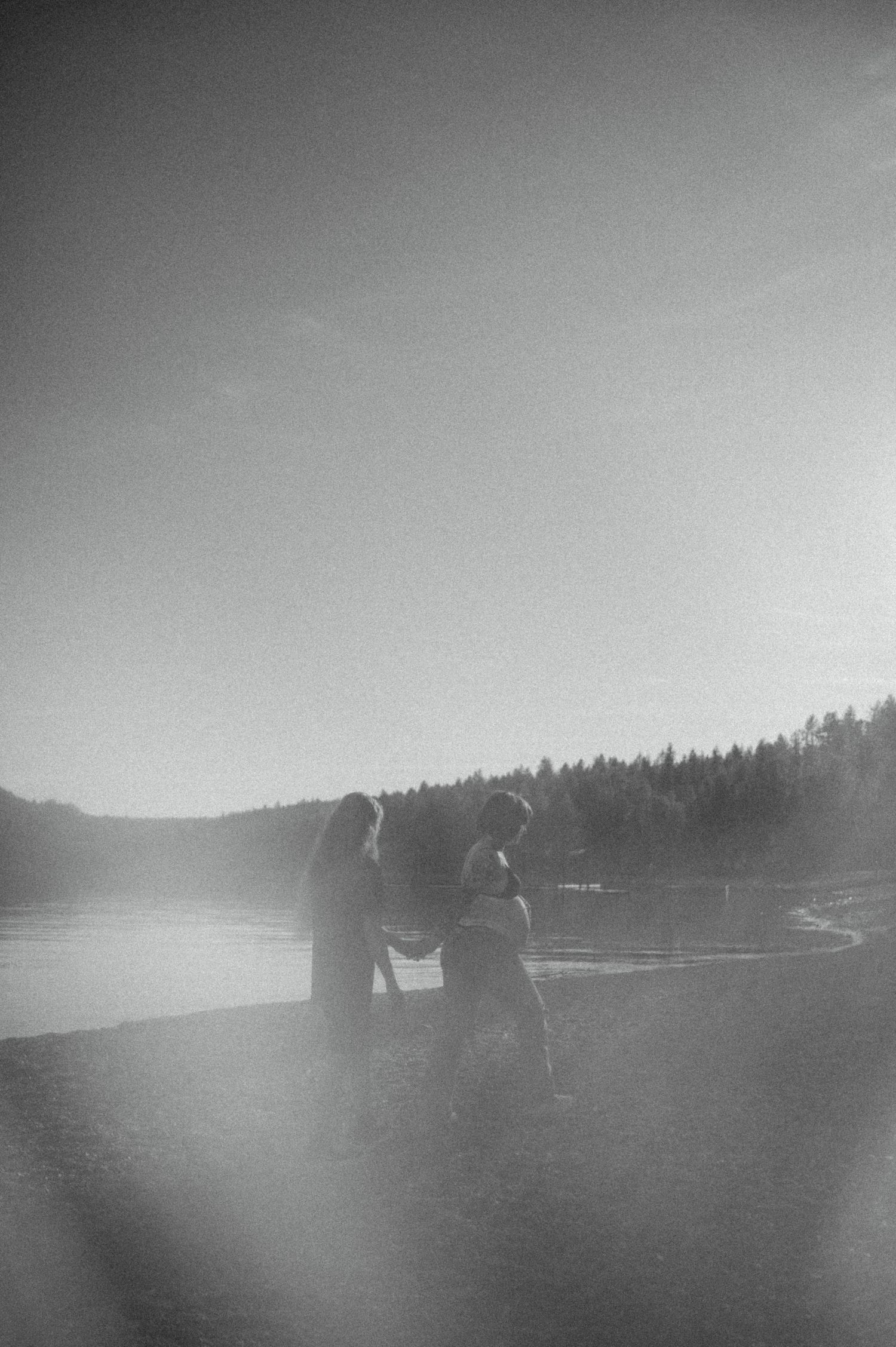 Two silhouetted figures stand at the edge of a misty lake surrounded by forested hills in black and white photography.