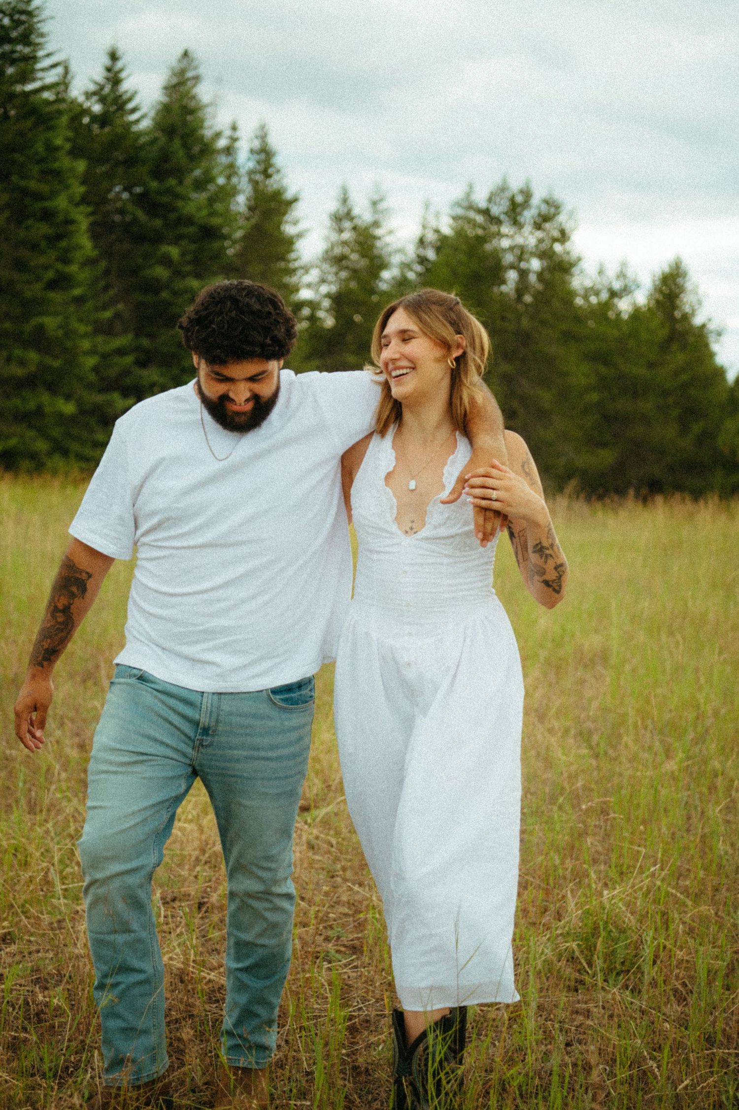 A couple shares a tender moment while standing together in a grassy meadow surrounded by evergreen trees.