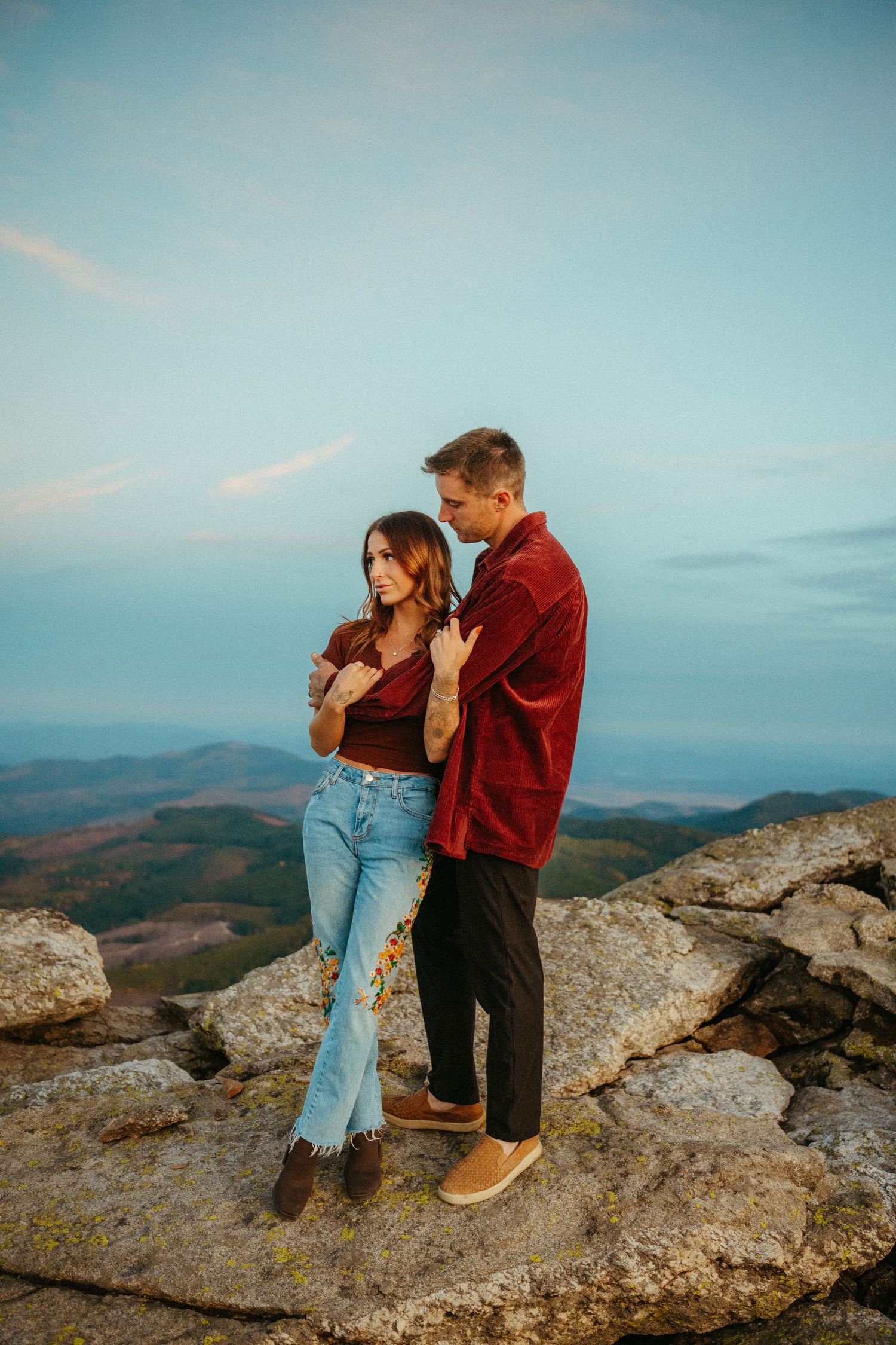 A couple stands together on a rocky mountain overlook during sunset, wearing casual clothes against a blue sky backdrop.