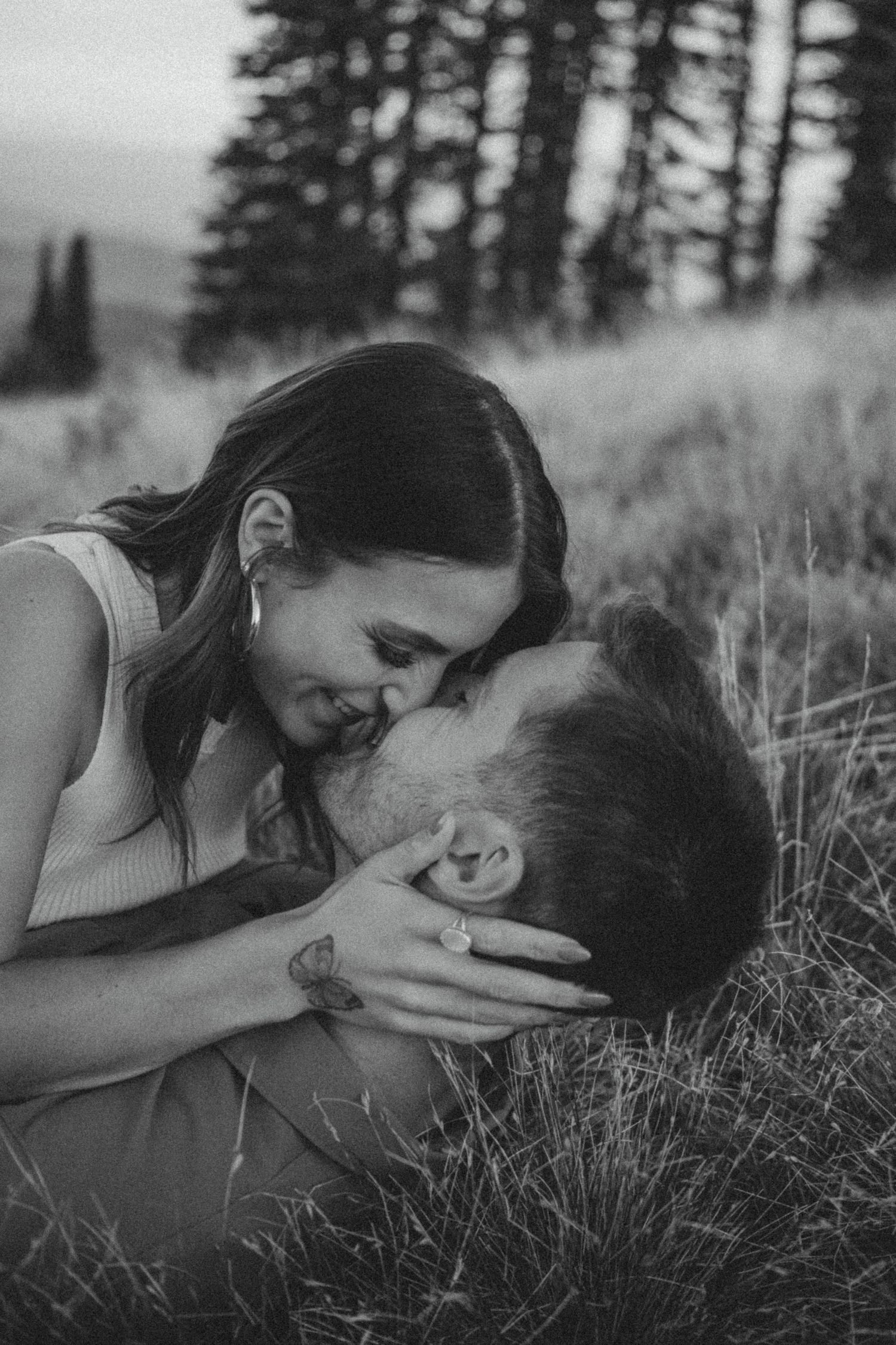 A tender black and white romantic moment captured between a couple laying in tall grass with pine trees in the background.