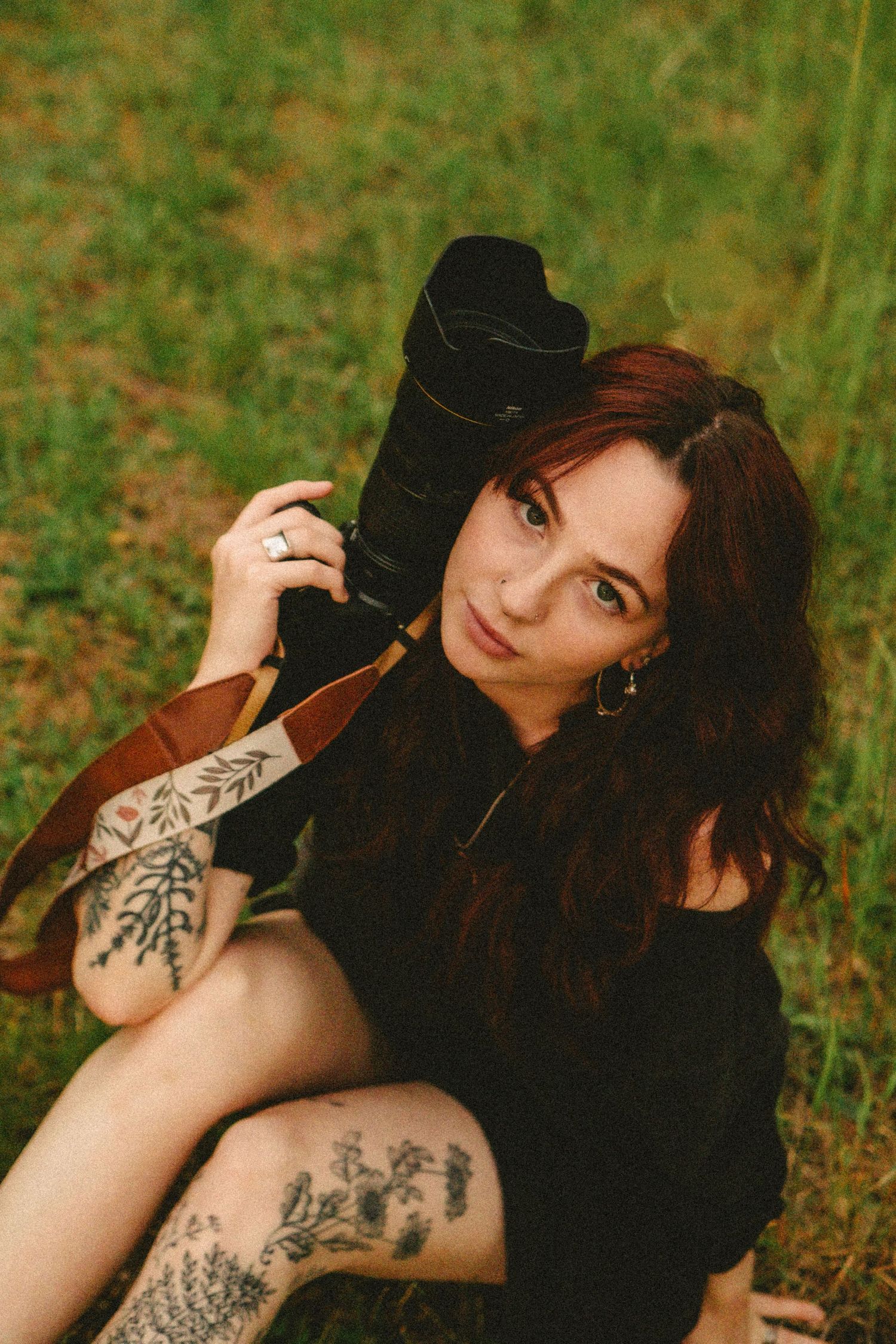 Portrait of someone with dark hair wearing black clothing sitting on grass, showing artistic tattoos in warm lighting.
