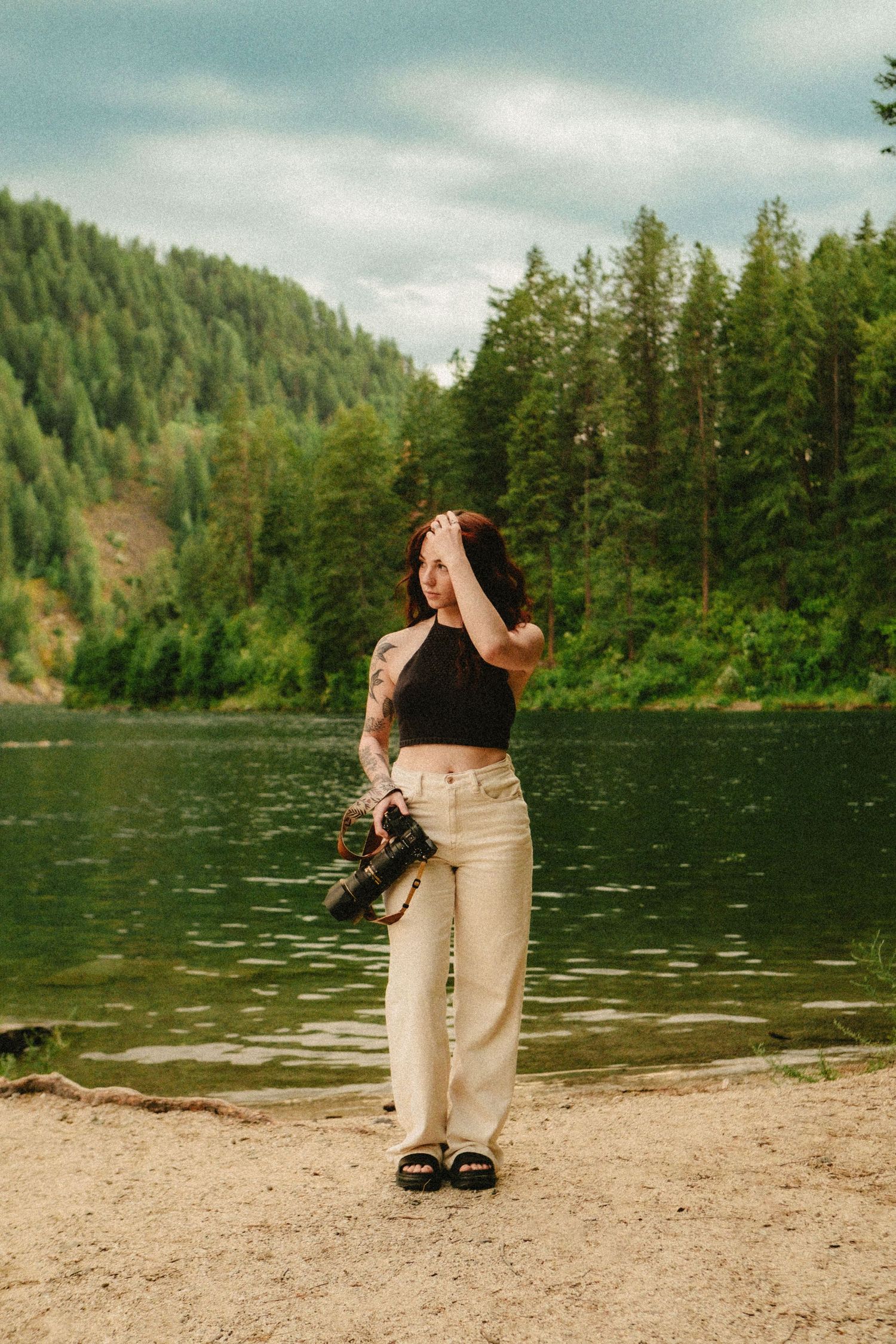 A figure stands at the shore of a serene lake surrounded by evergreen forests on a cloudy day.