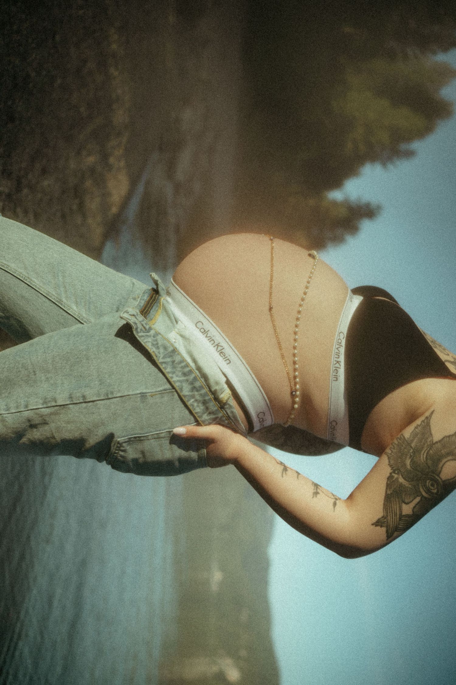 Artistic photo showing tattoo on arm with vintage jeans and beige top against hazy blue sky background.
