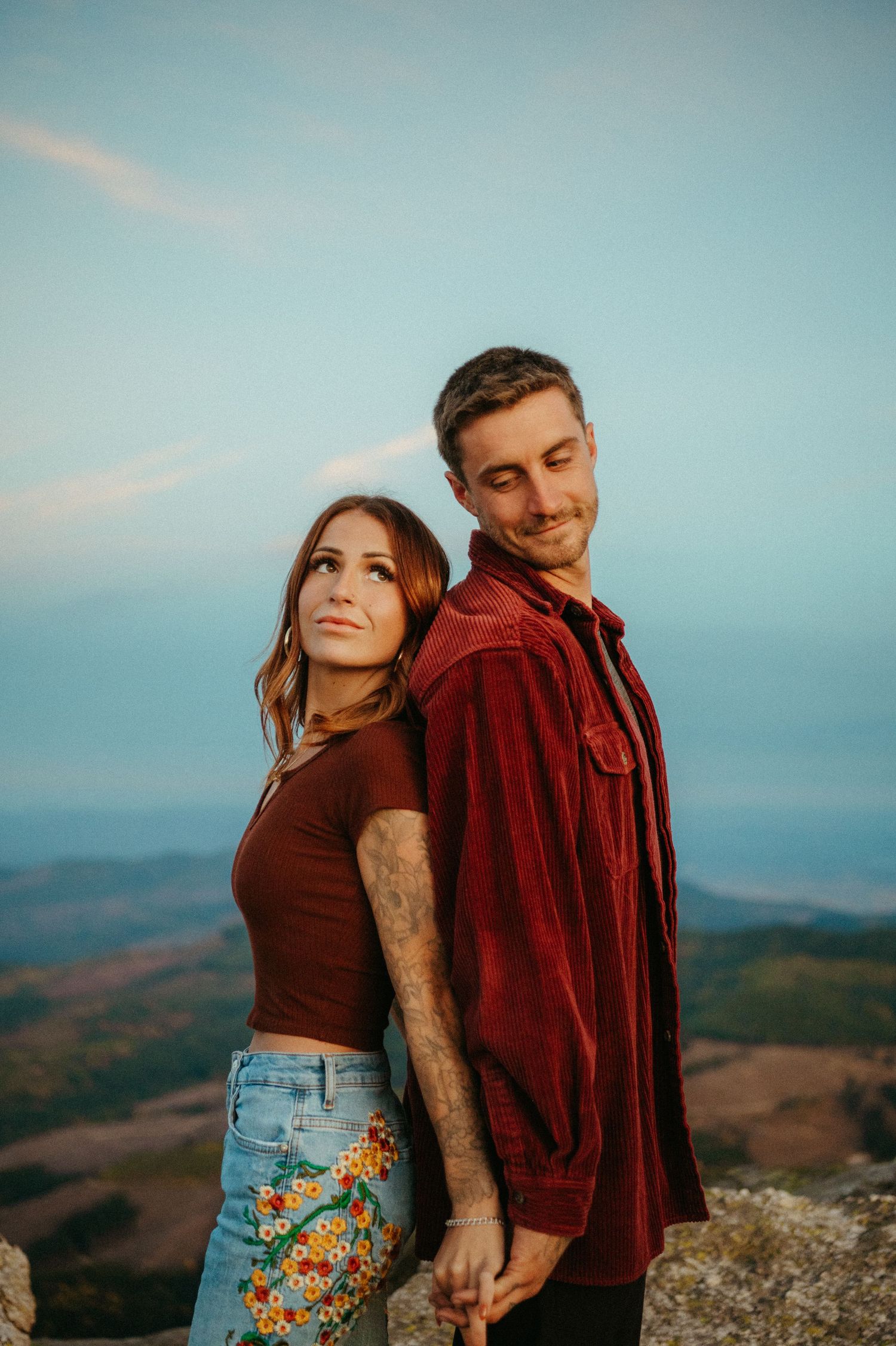 A couple stands close together on a mountain overlook at sunset wearing burgundy and brown casual attire.