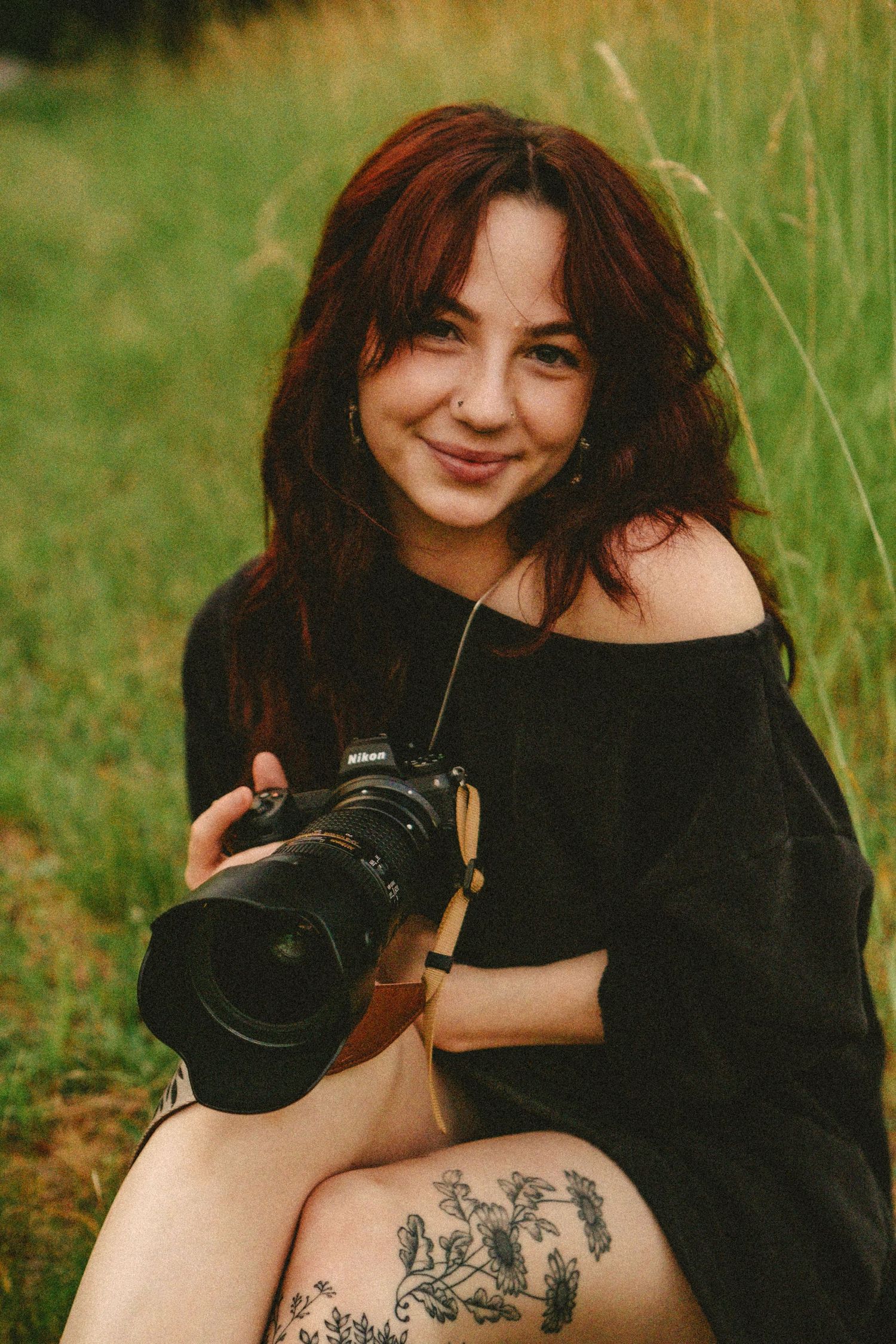 A photographer in a black off-shoulder dress sits in tall grass holding a professional camera with a leg tattoo visible.