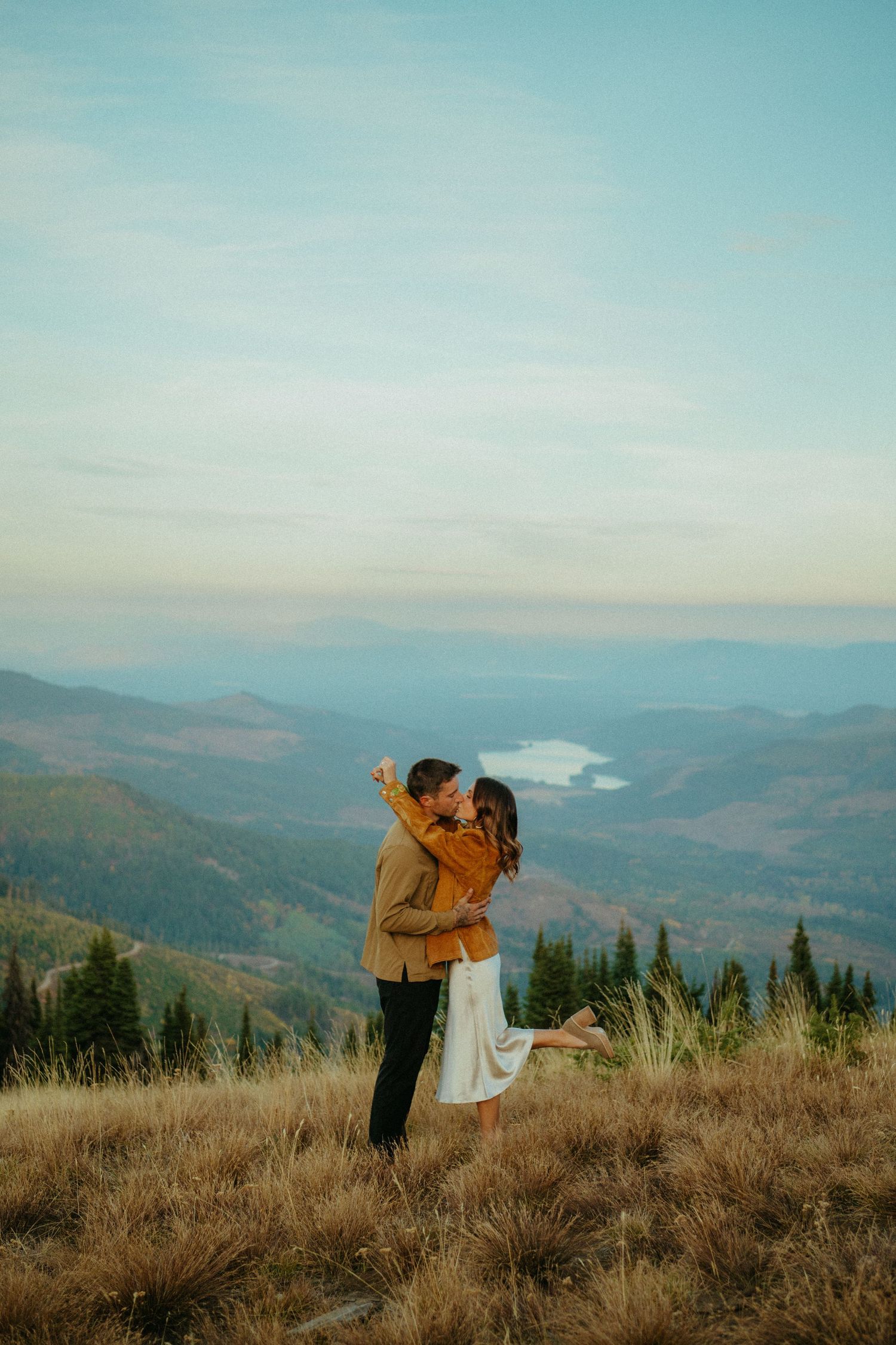 A romantic couple embraces on a mountain summit overlooking a scenic valley during golden hour.