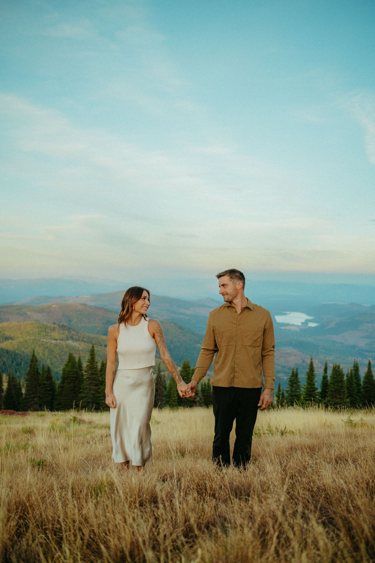 A couple holding hands while standing in a mountain meadow during sunset overlooking scenic mountain vistas and a lake.
