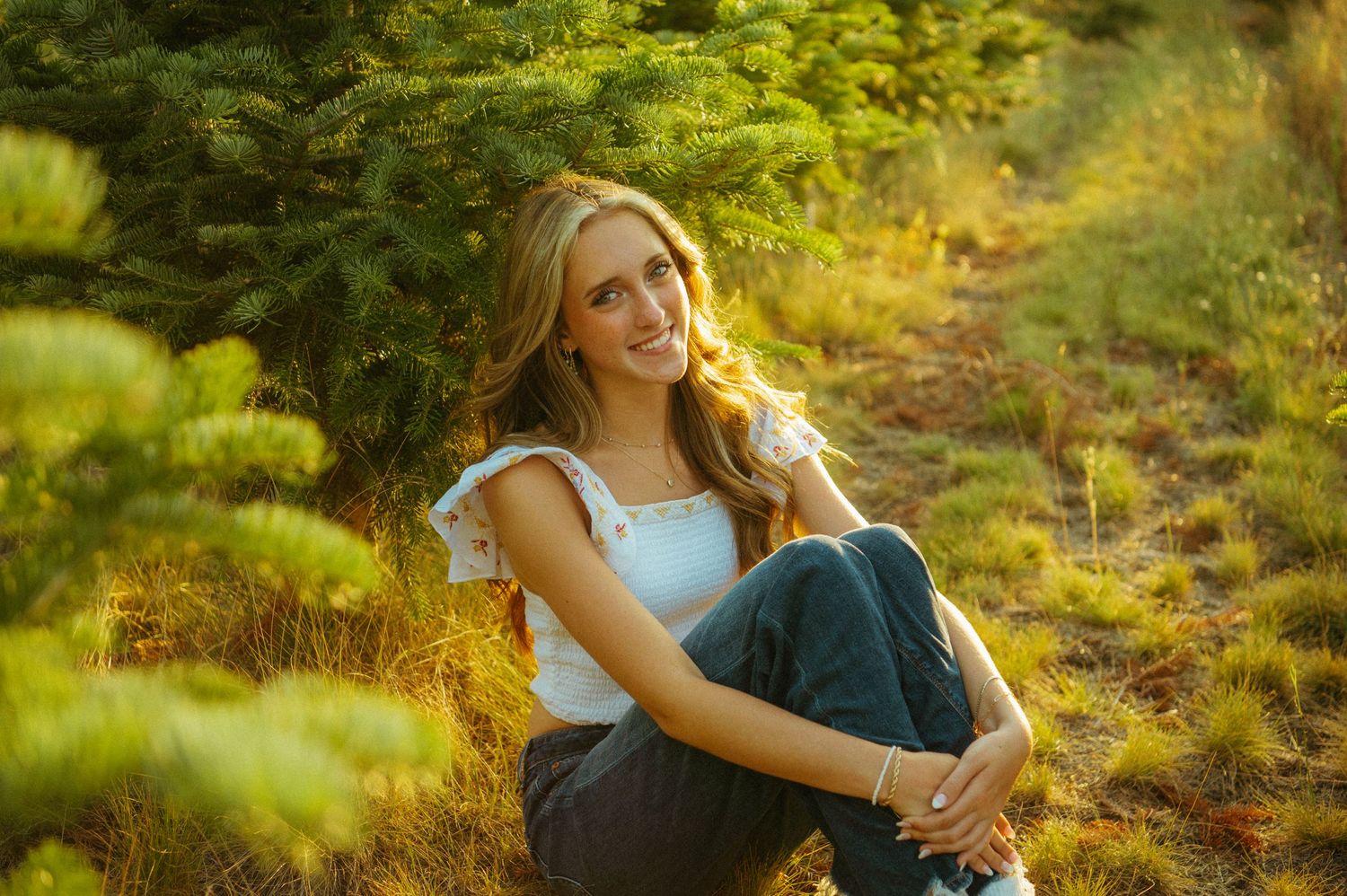 A person sits in a golden-lit forest wearing jeans and a light blue top, surrounded by lush green foliage.