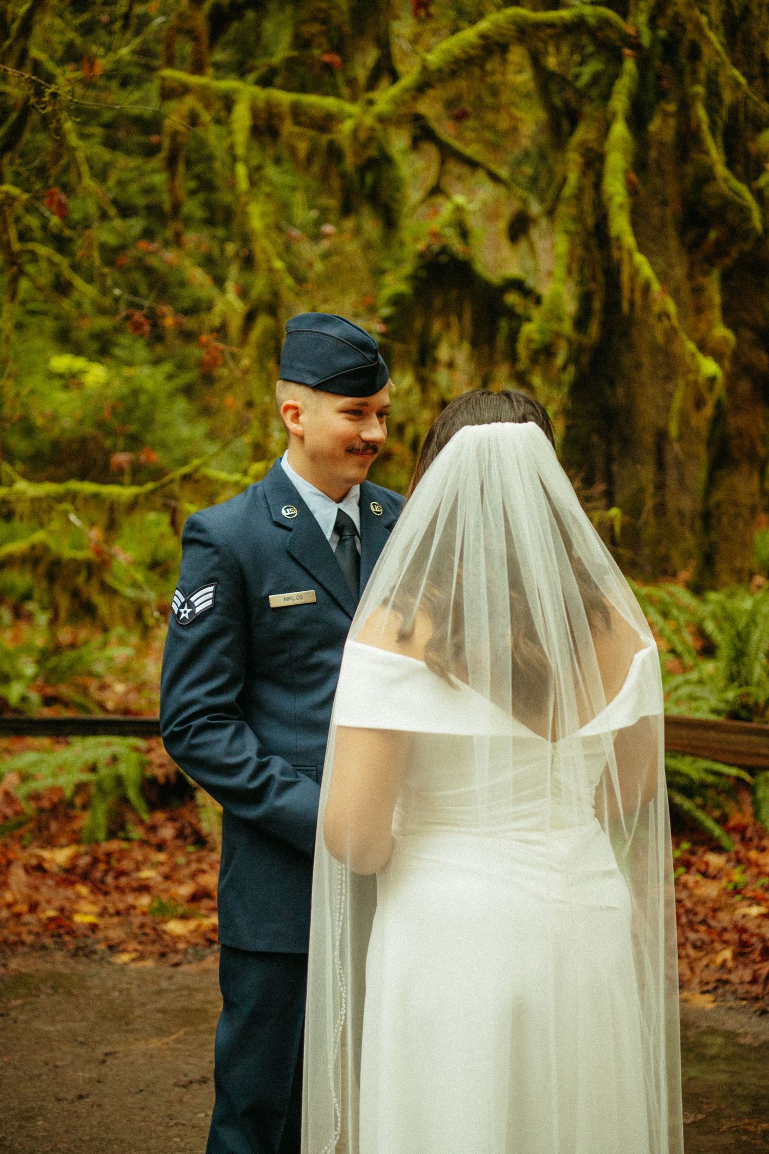 A military service member in dress blues and a bride in a flowing white gown and veil share a moment in a mossy forest.