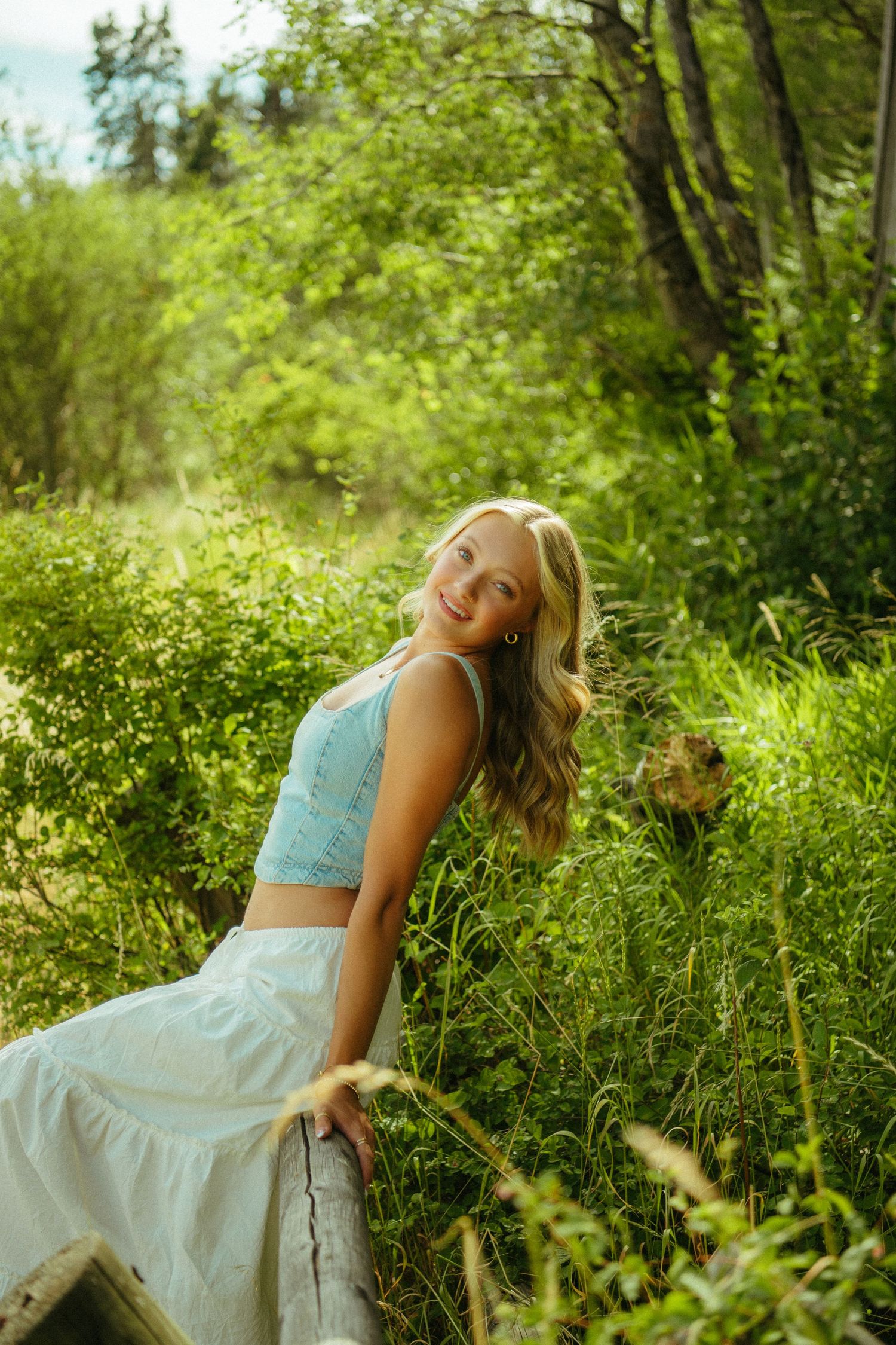 A person in a light blue dress sits gracefully among lush green foliage in a sunlit forest setting.