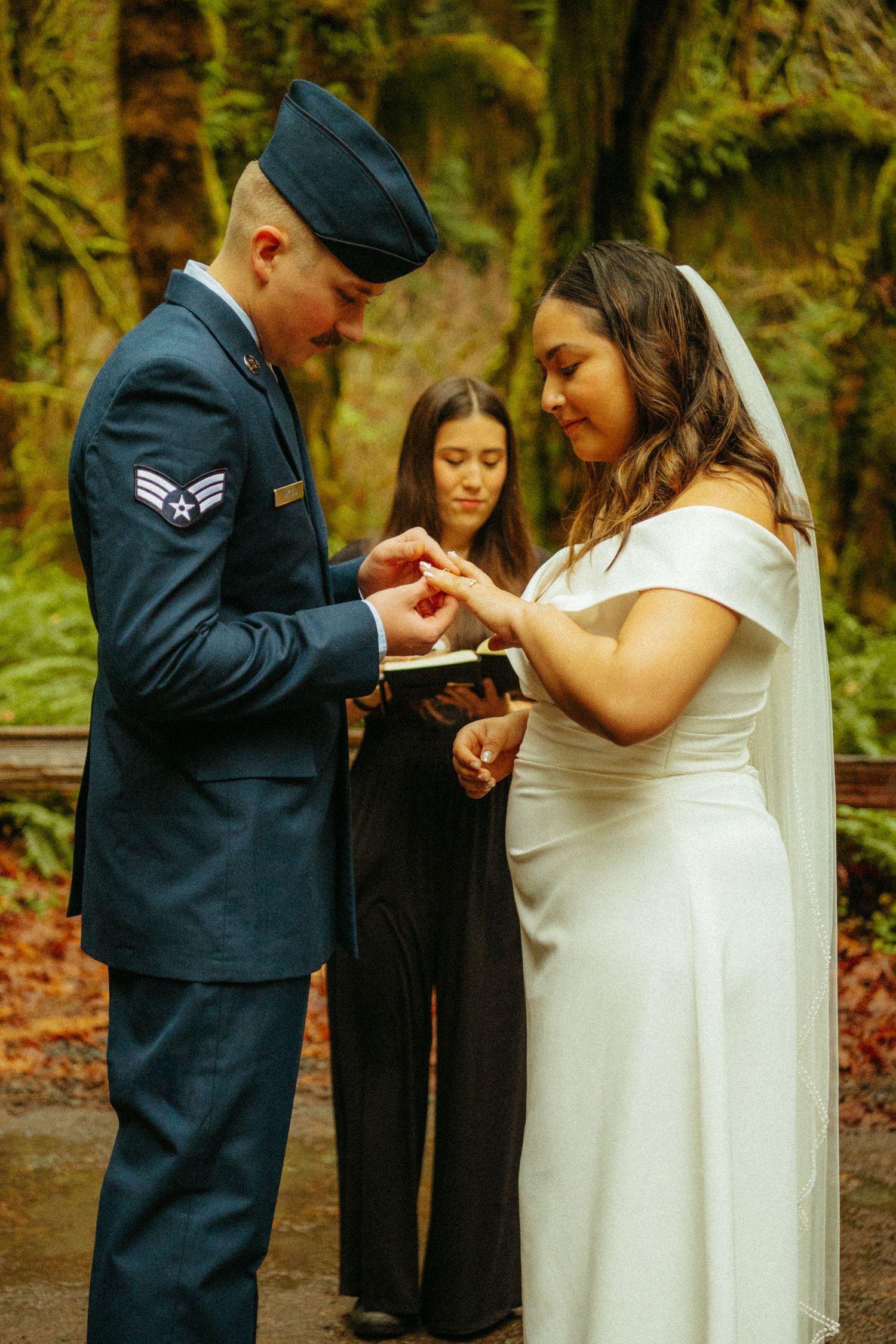 Military service member exchanges wedding rings with bride during outdoor autumn ceremony.