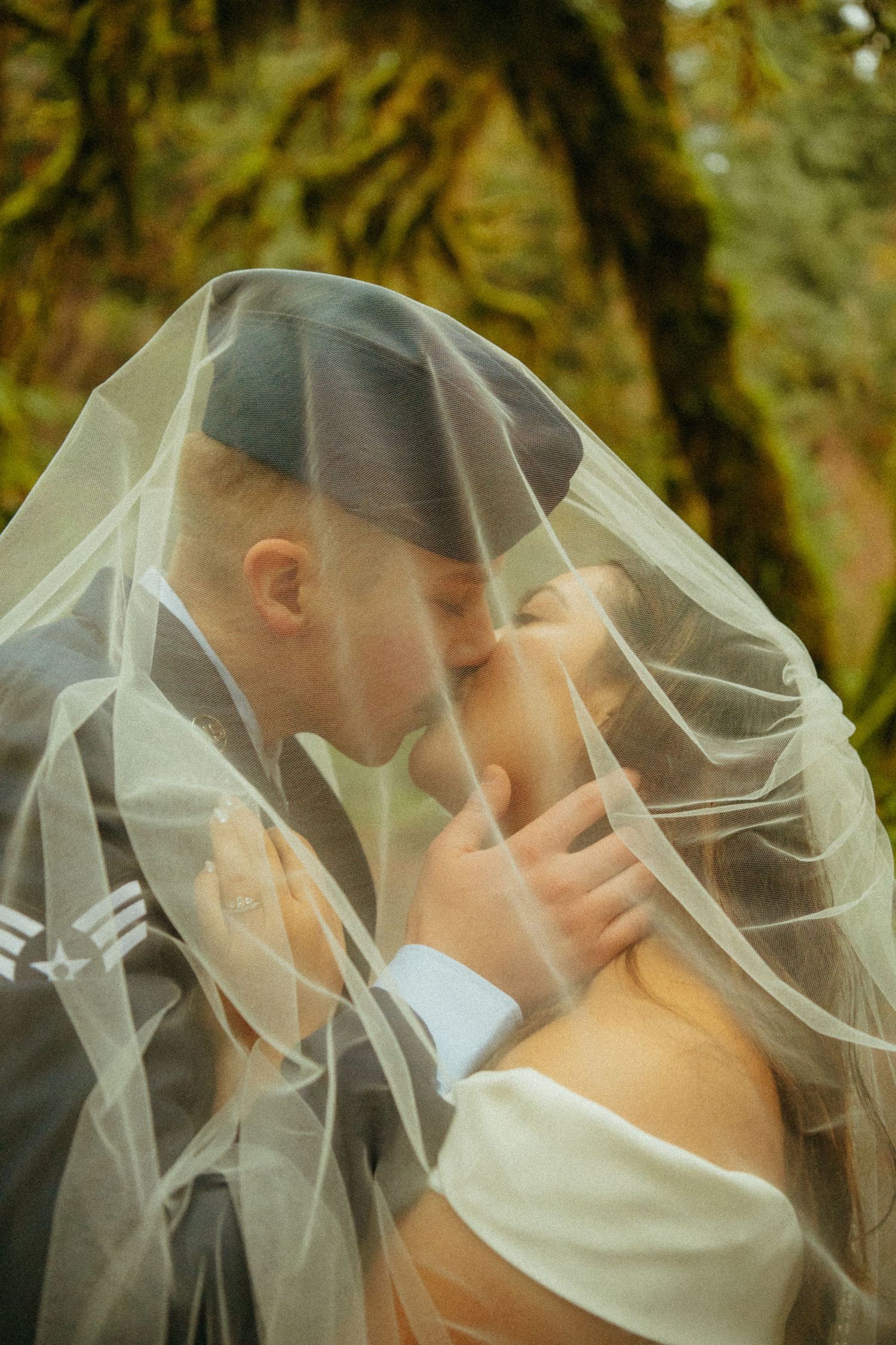 A romantic wedding moment captured under a delicate white veil in a dreamy forest setting with mossy trees.