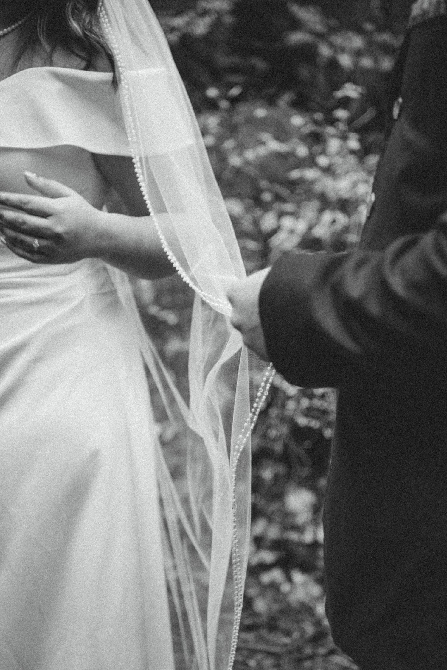 A close-up black and white photo of a delicate lace wedding veil and white wedding dress during an outdoor ceremony.