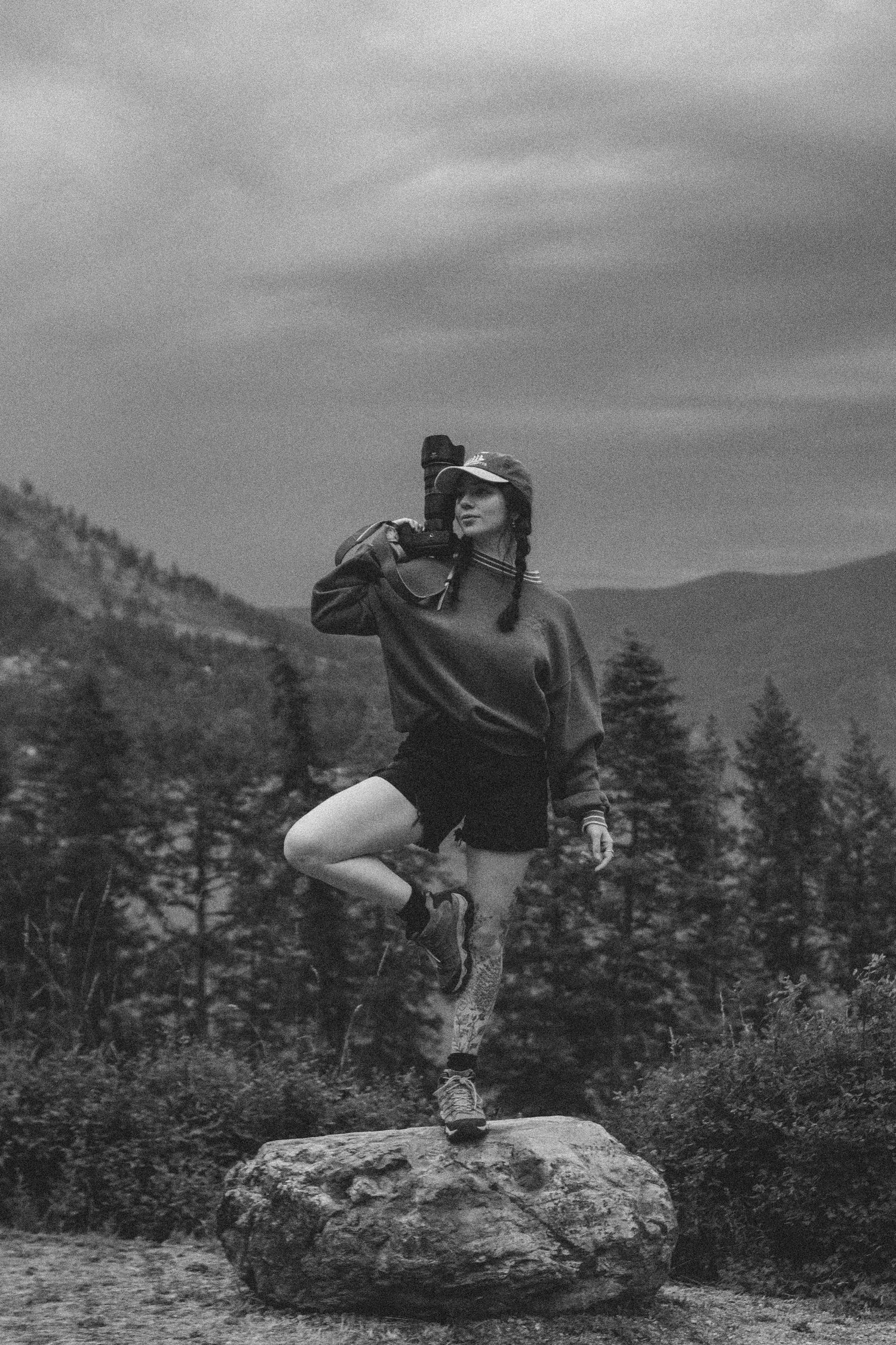 A person strikes a dynamic dance pose while balanced on a rock against a mountainous landscape in black and white.
