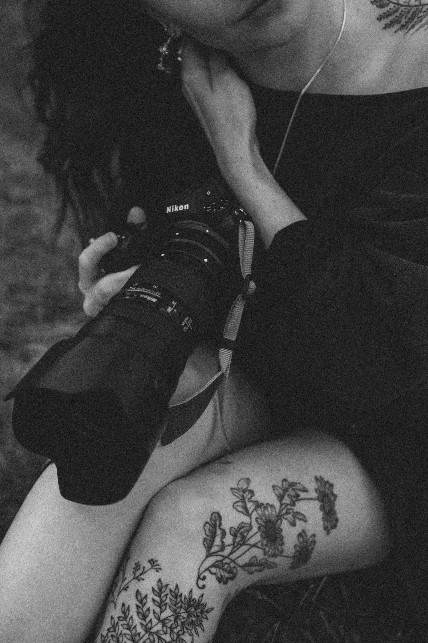 Black and white photo of photographer with Nikon camera and floral leg tattoo sitting outdoors.