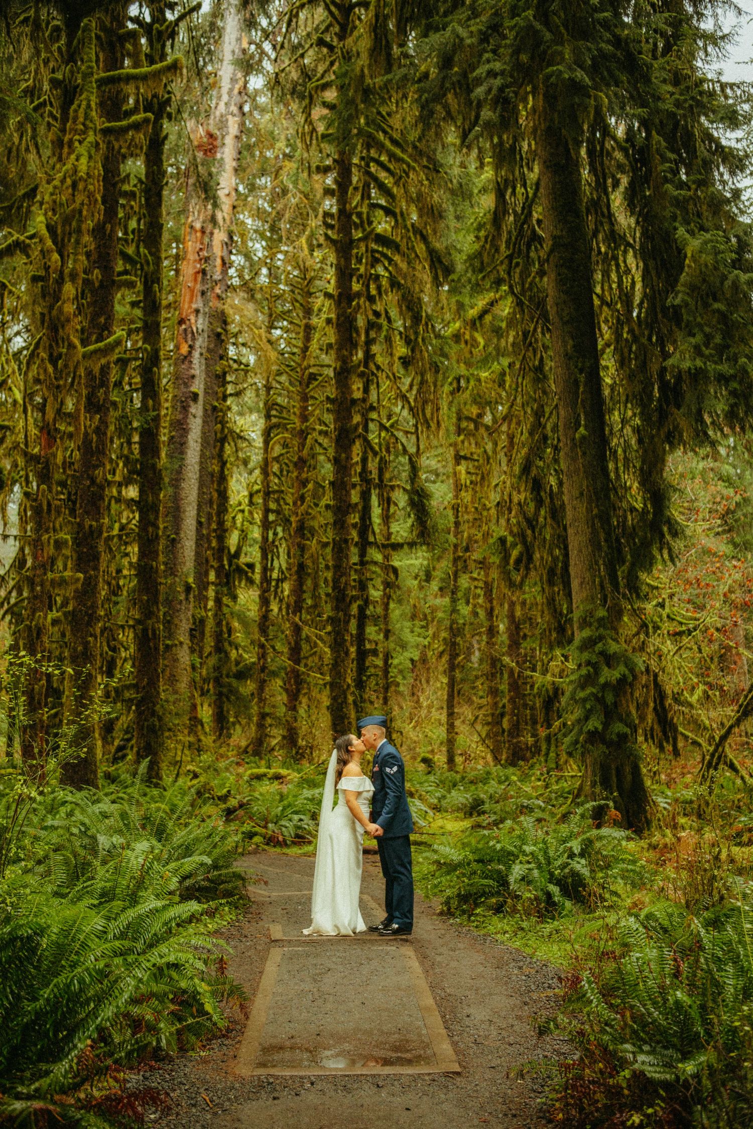 A couple in wedding attire stands together on a forest trail surrounded by towering moss-covered trees and ferns.