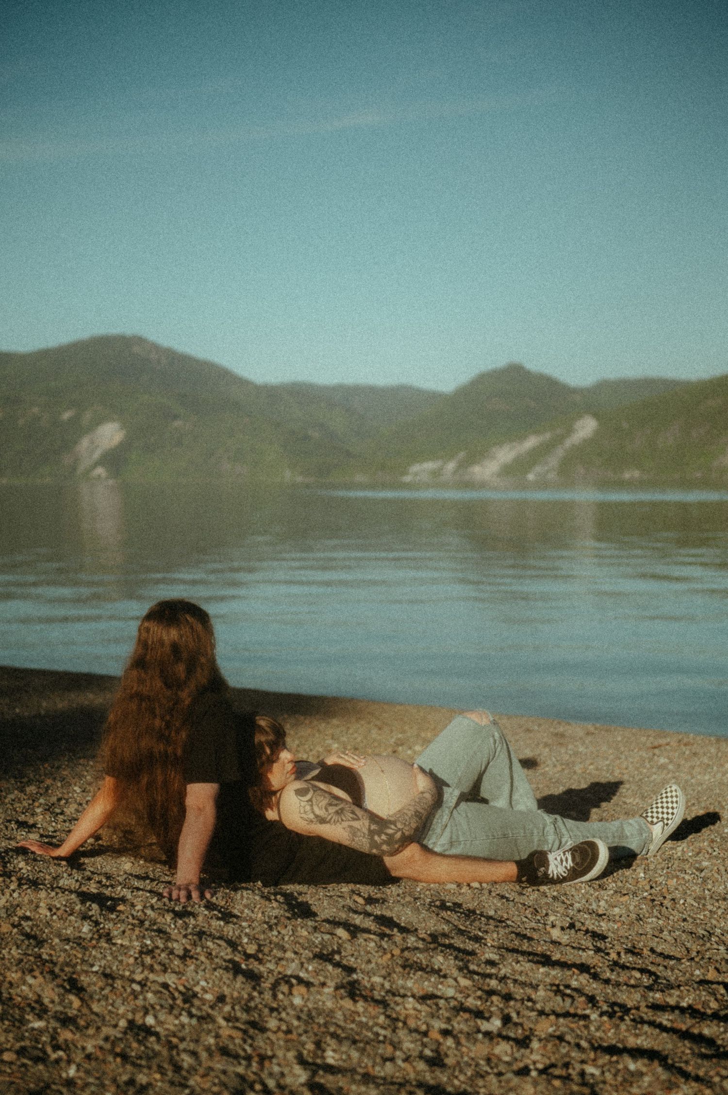 Person relaxing on sandy shore beside mountain lake under blue sky on sunny day.