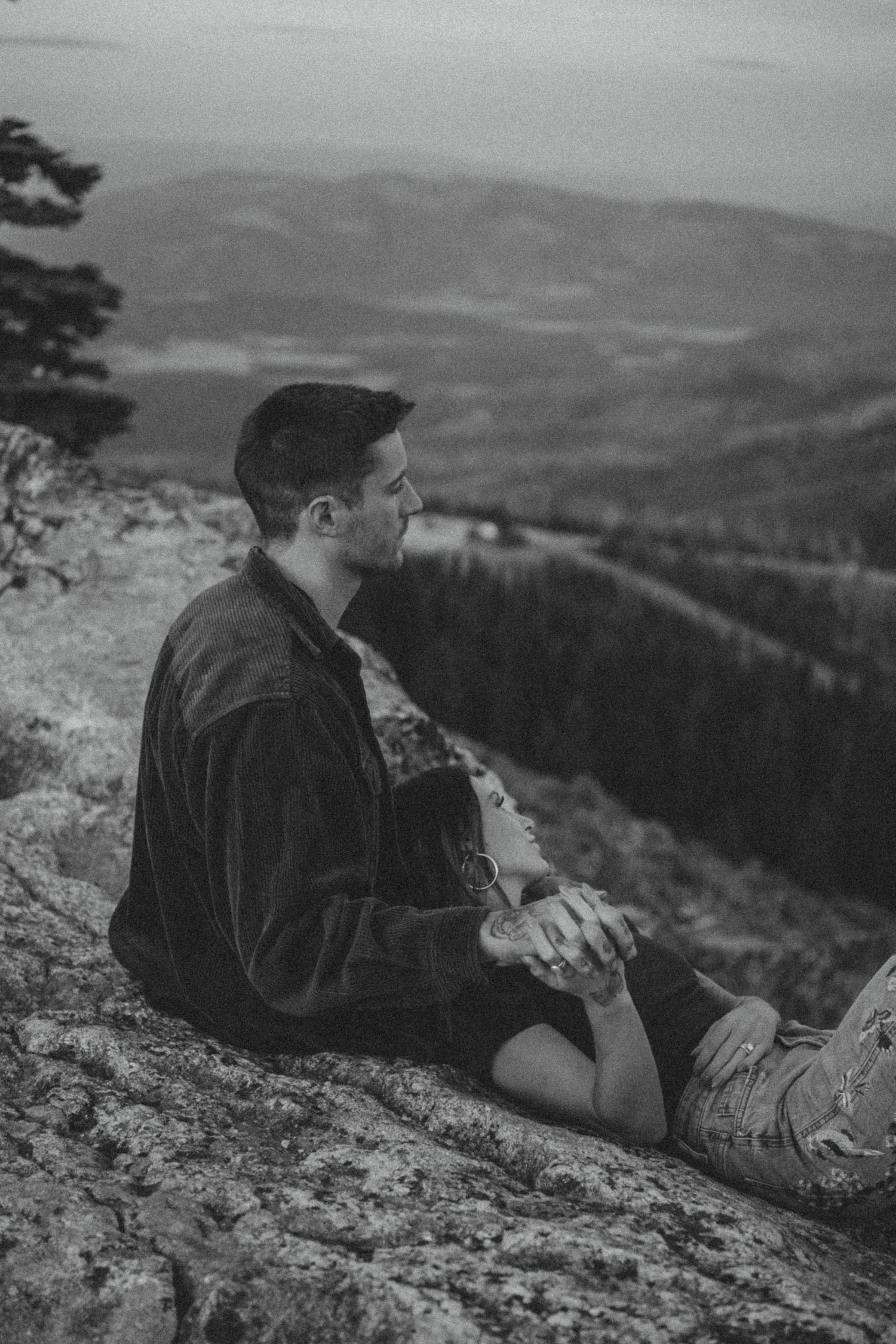 A black and white photograph of someone sitting on a mountain ledge overlooking a vast valley landscape below.