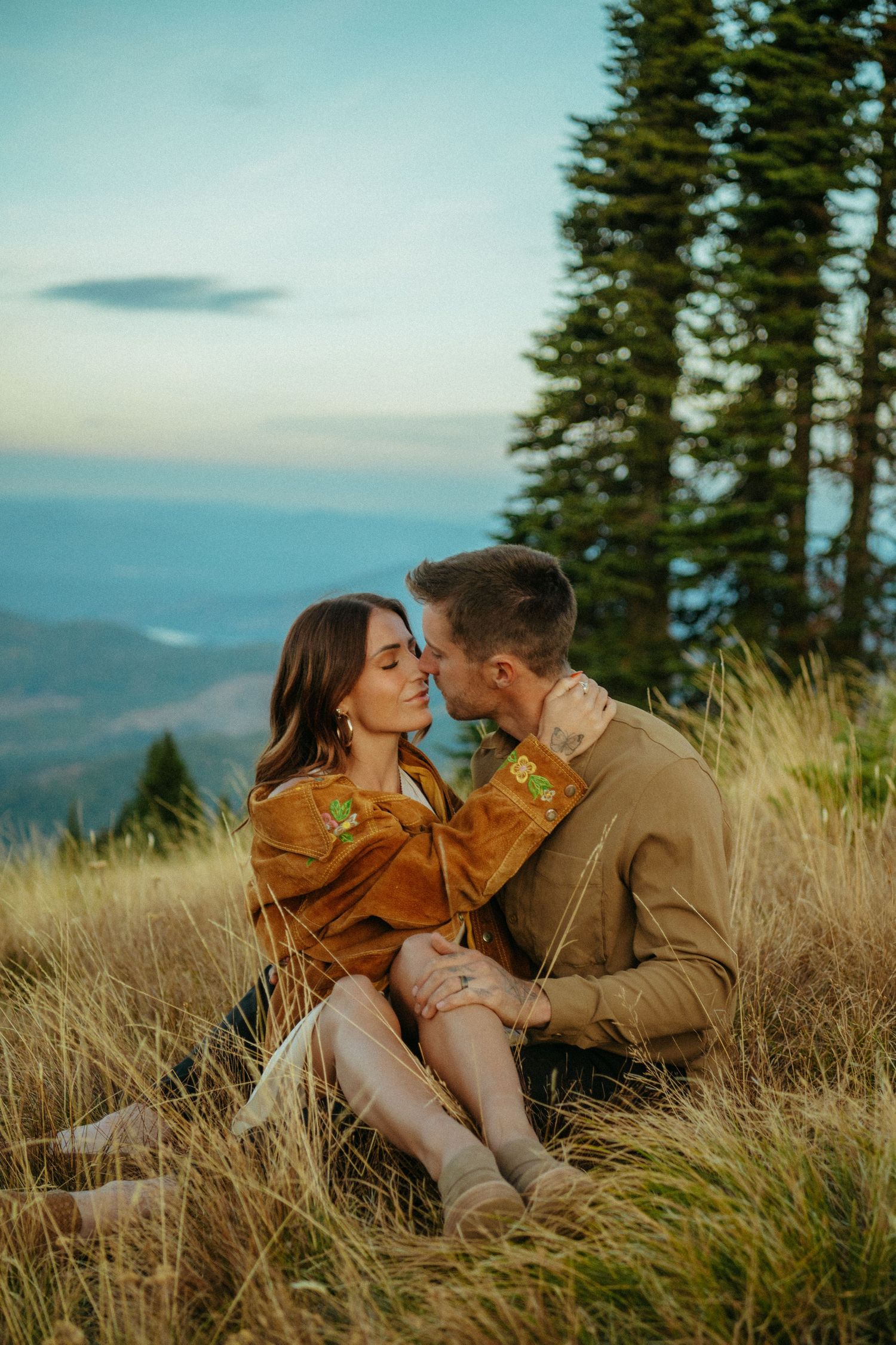 A couple sits together in tall grass on a mountainside at sunset with evergreen trees and mountain views in the background.