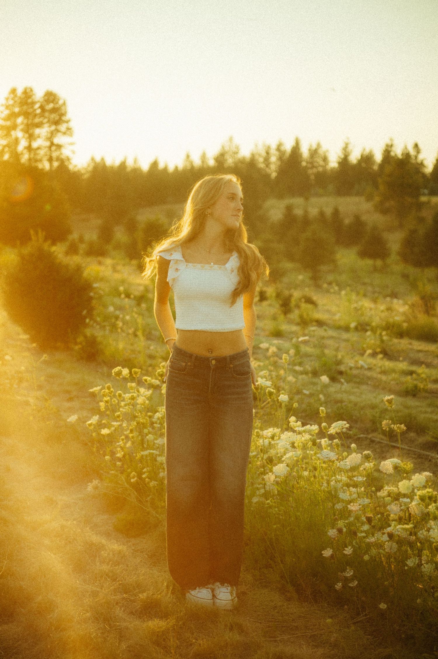 Figure in a white crop top and wide-leg jeans stands in a wildflower field at golden hour with sunlit trees in background.