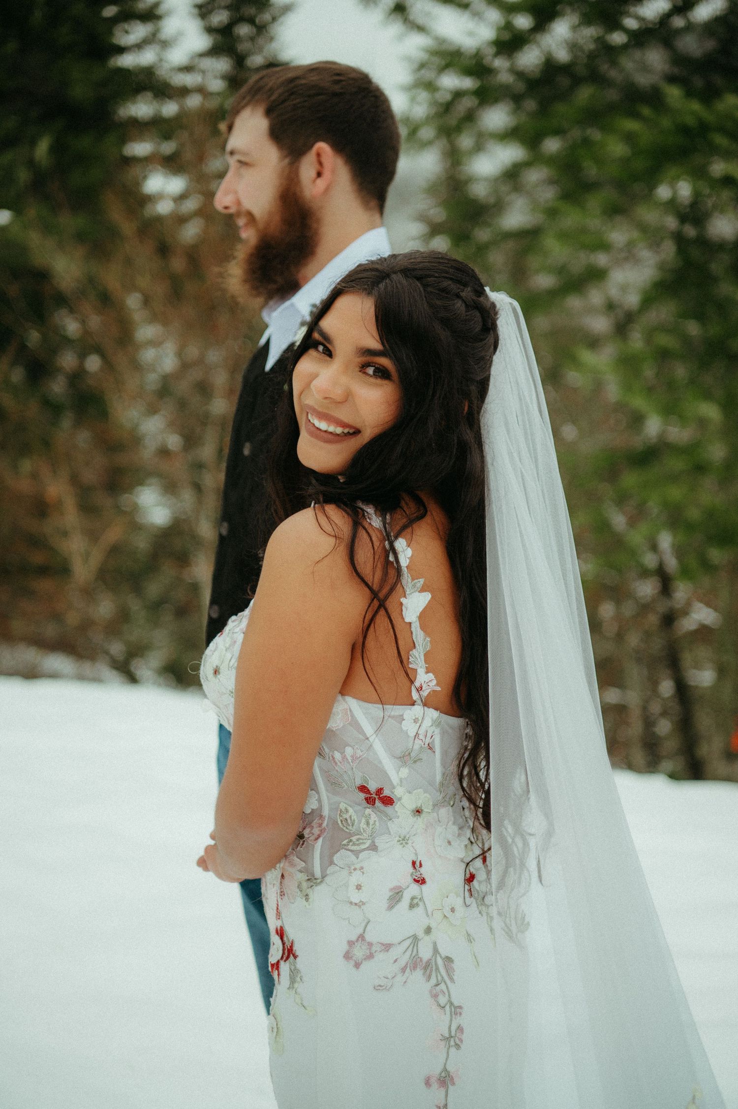 A winter wedding scene shows a couple in formal attire embracing in a snow-covered forest setting.