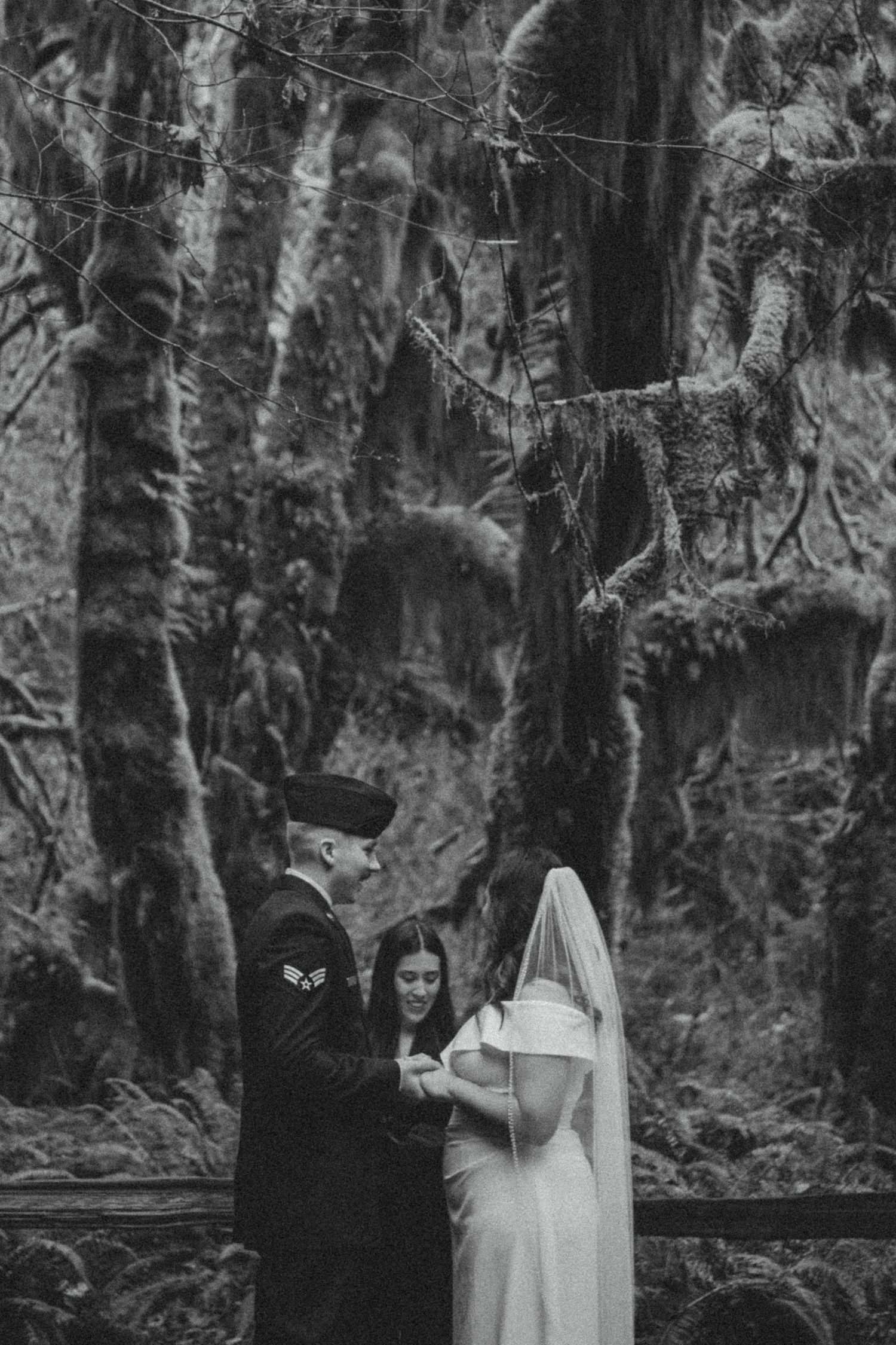 Black and white photograph of a couple in wedding attire standing in a mossy forest setting.