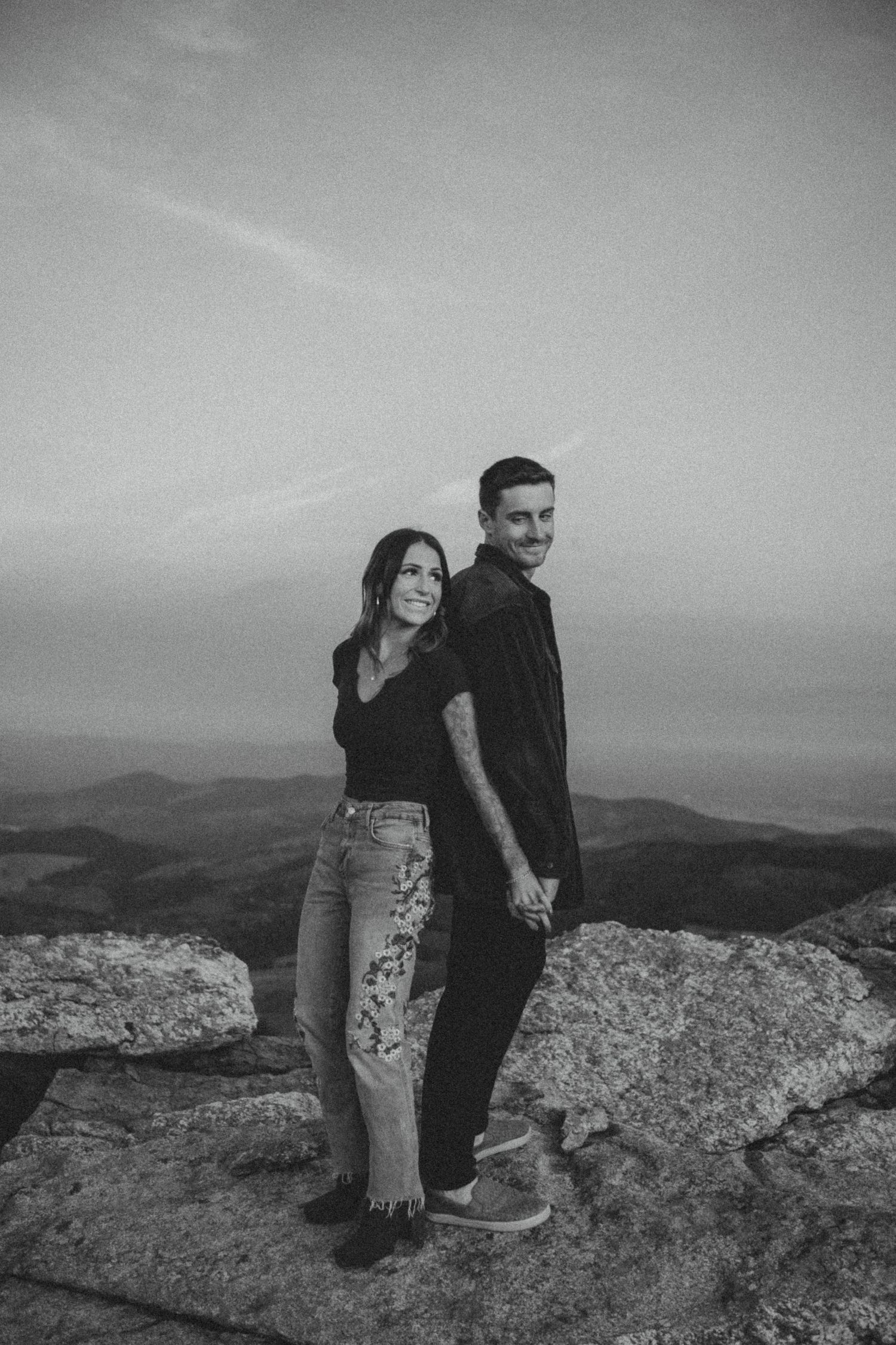 A couple stands together on a rocky mountain overlook in black and white, with rolling hills visible in the distance.