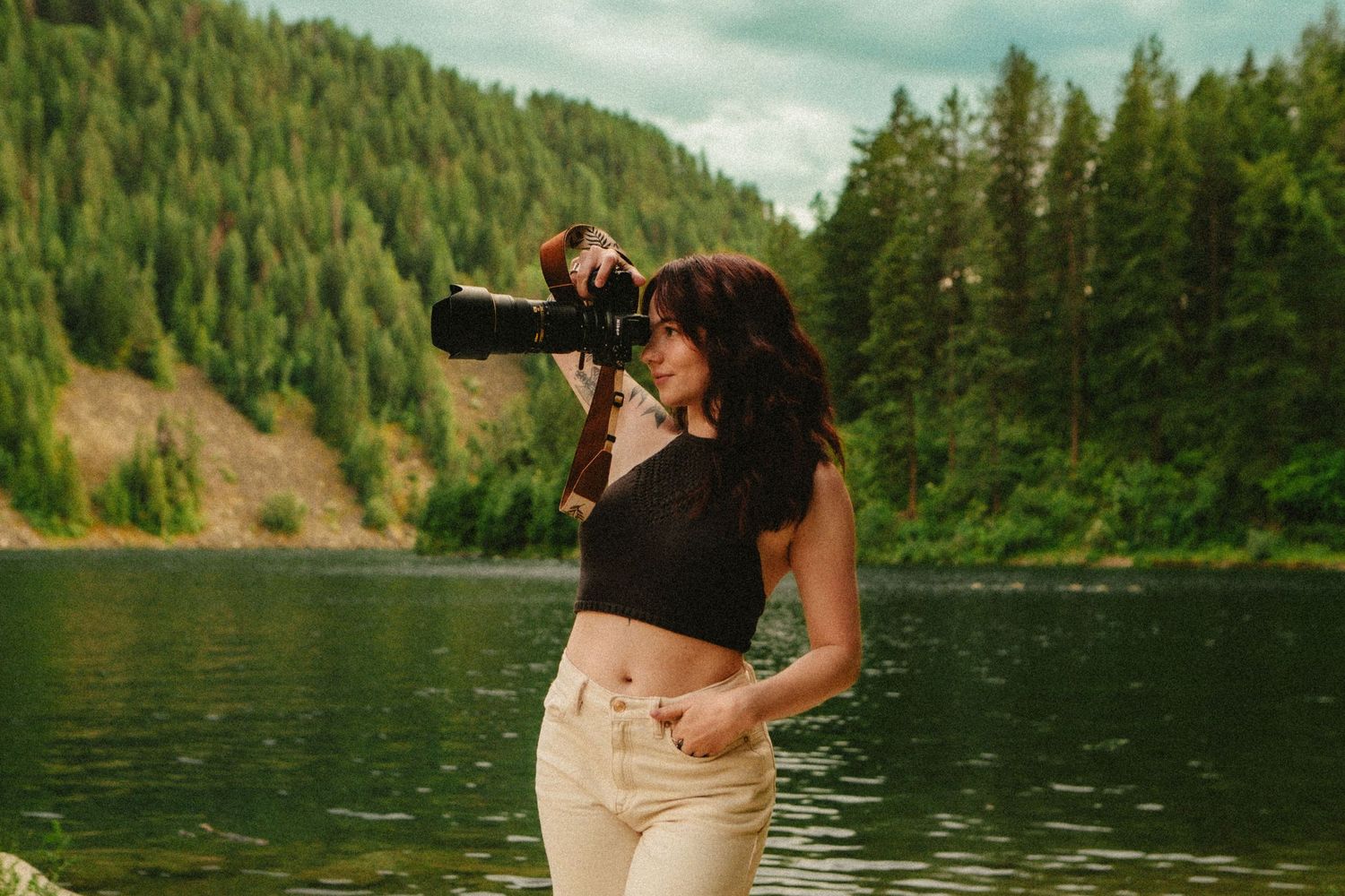 Someone takes a picture from a camera while standing by a serene lake surrounded by evergreen forest slopes.