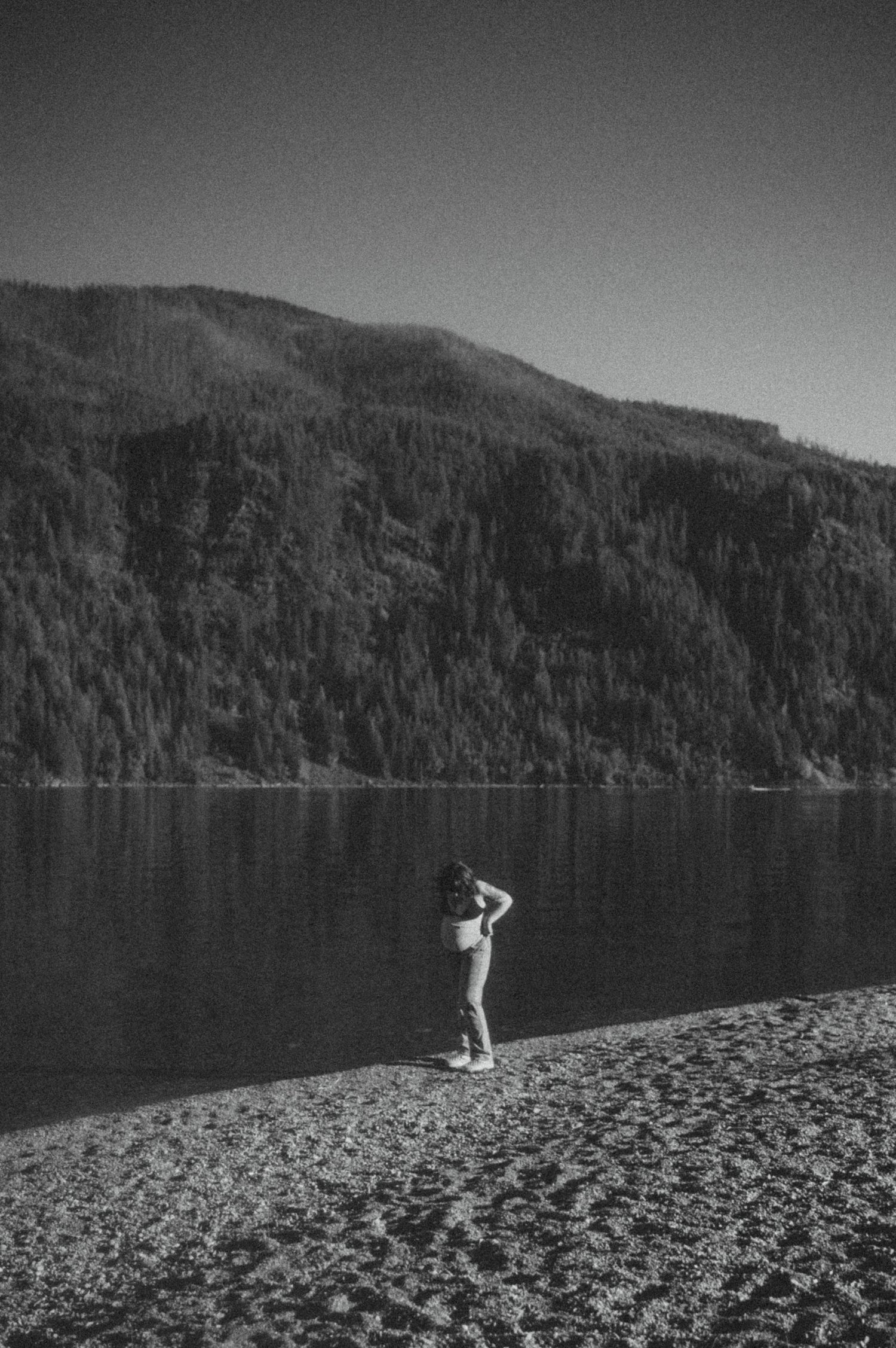 A lone figure stands on a rocky beach beside a serene mountain lake in black and white photography.