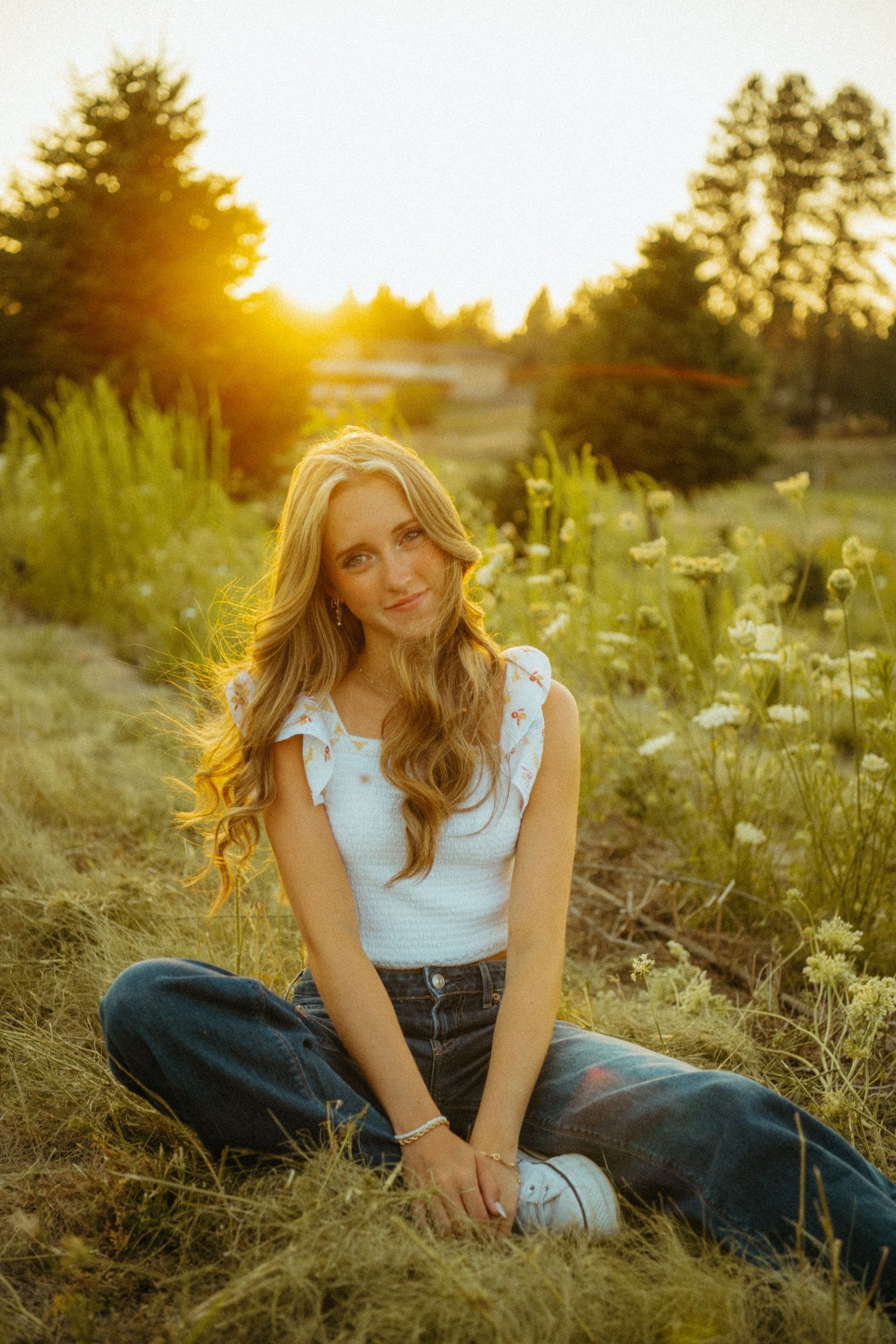 A golden sunset illuminates a meadow with tall grass while a woman in blue jeans and white top sits casually outdoors.