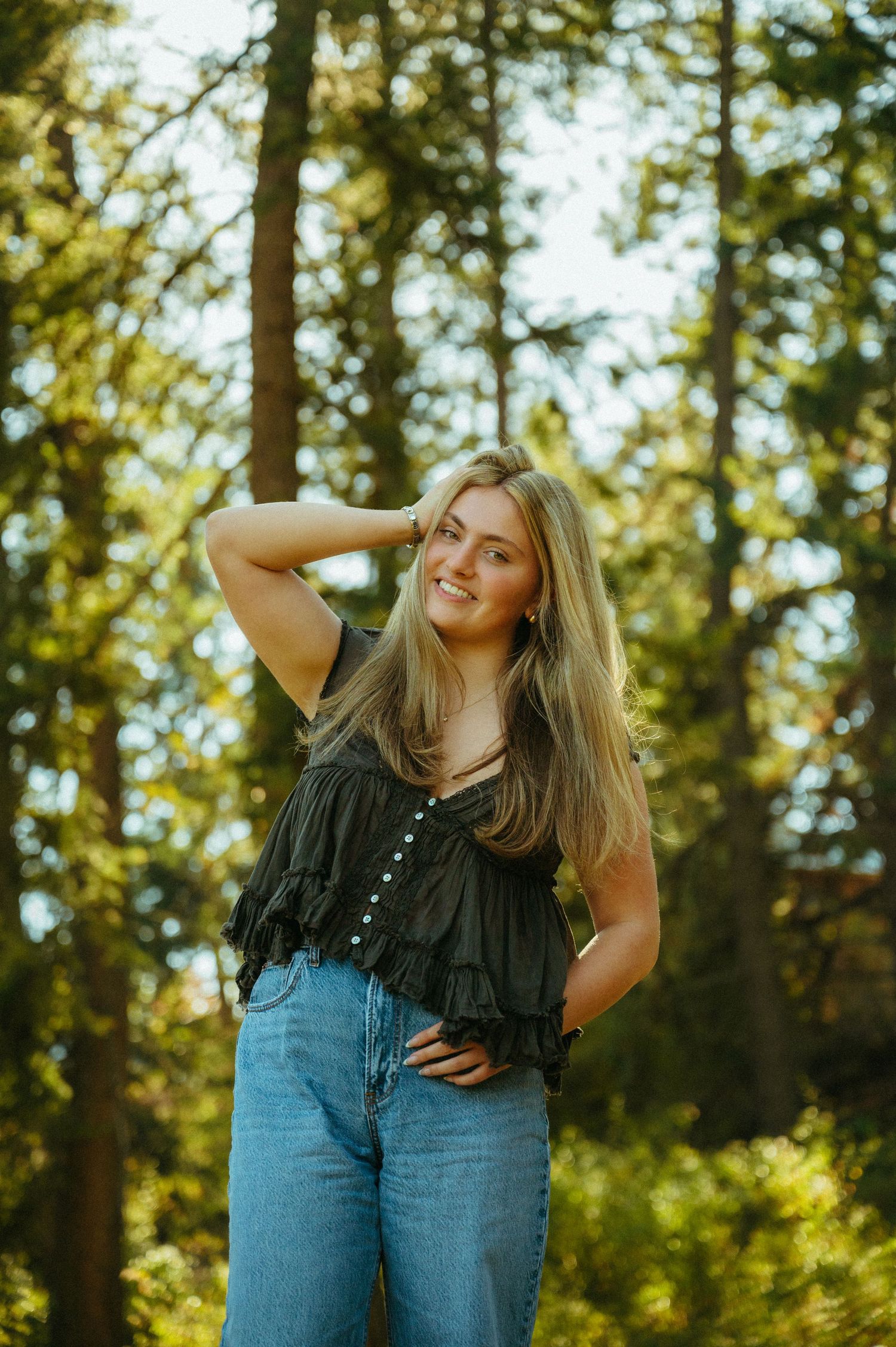 A woman in a black ruffled top and jeans poses playfully in a sun-dappled forest setting.