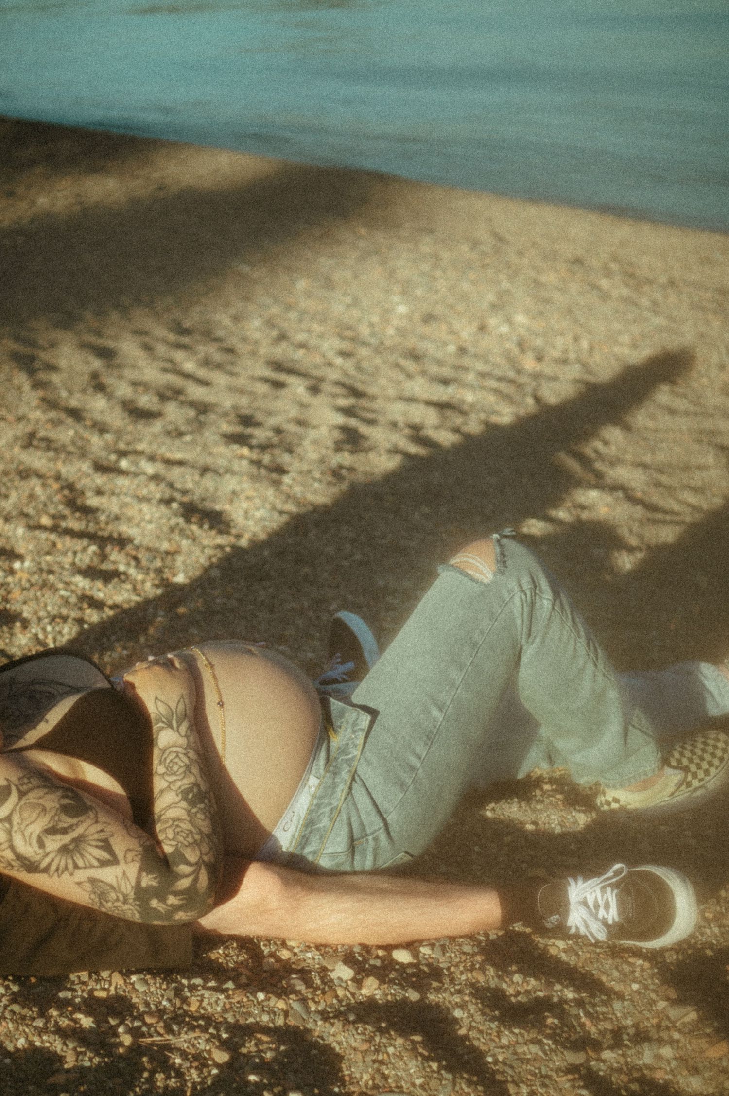 Person relaxing on sandy beach wearing blue jeans and sneakers with turquoise water in background.