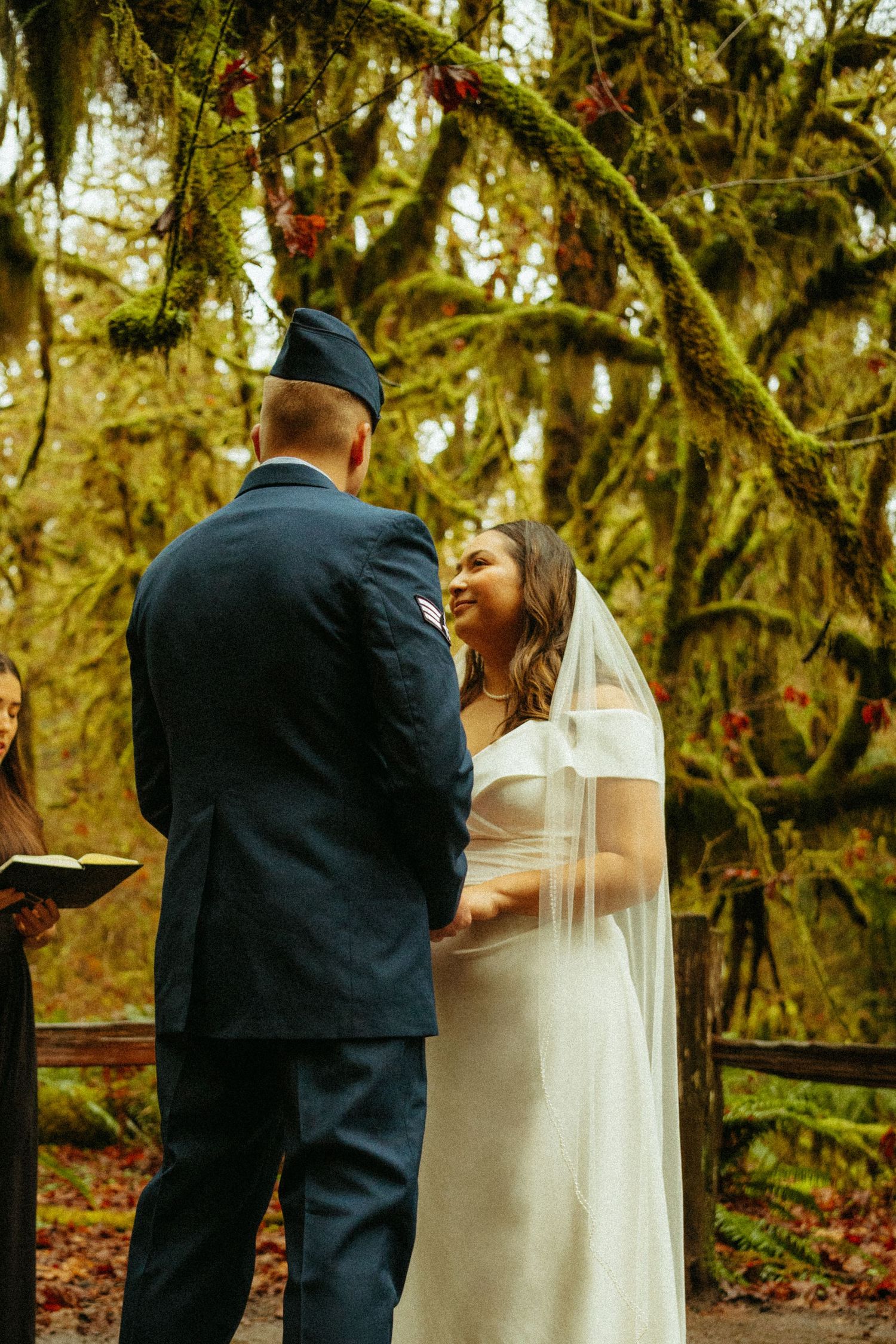 A military wedding ceremony takes place in a mossy forest with green branches hanging overhead.