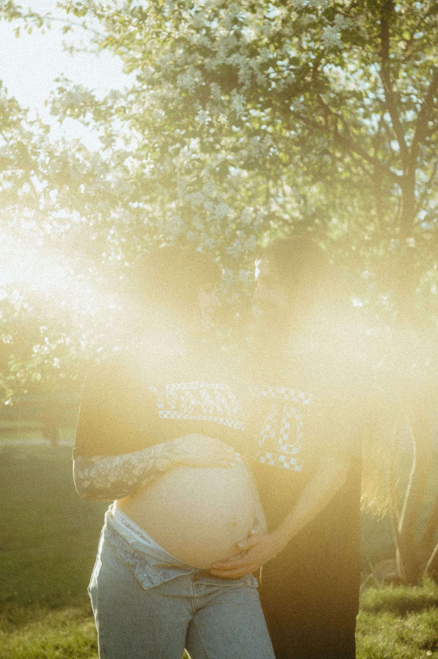 A silhouetted maternity photo taken at sunset with sunlight filtering through trees in the background.