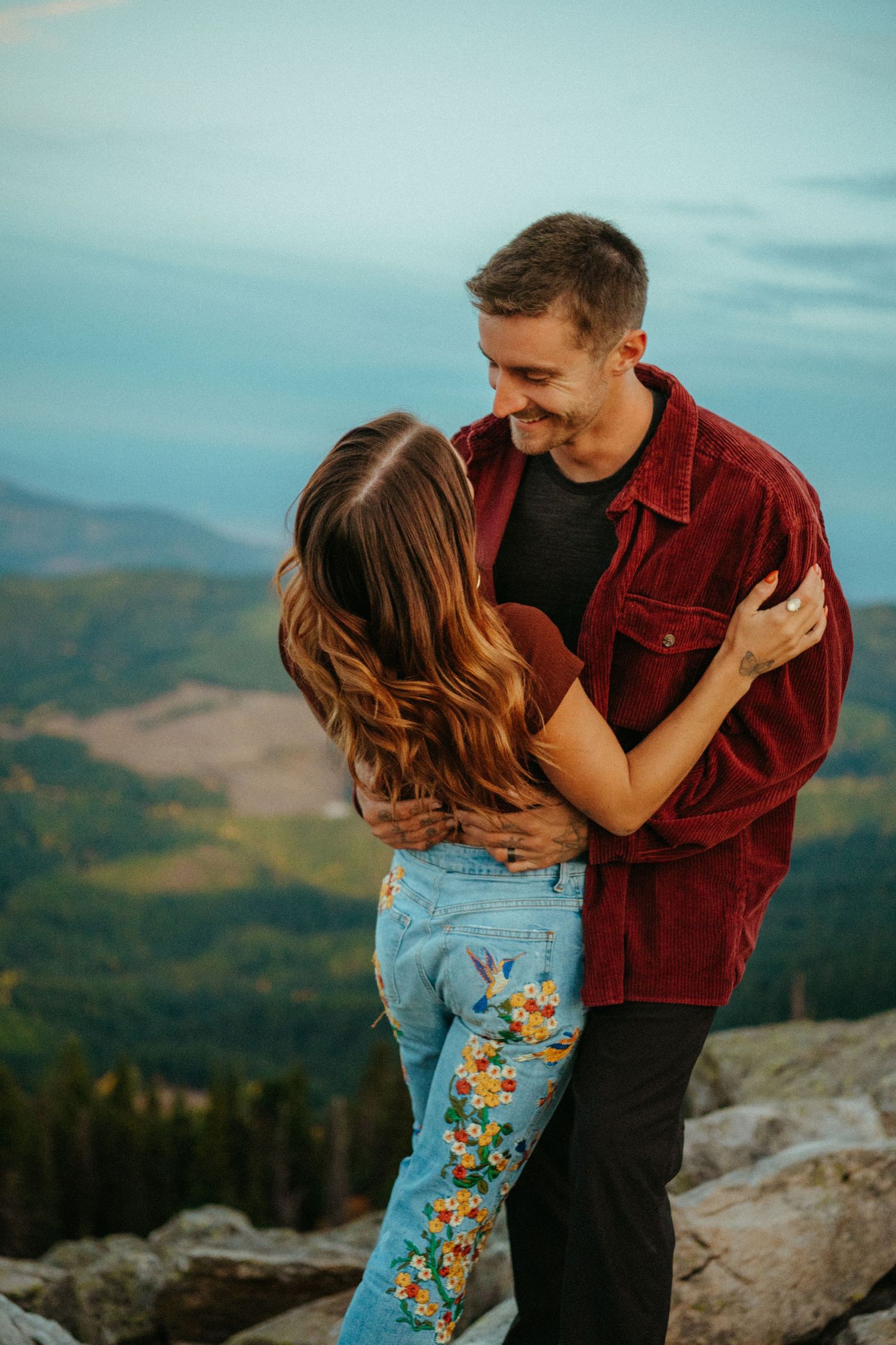 A couple embraces on a mountain overlook during sunset while wearing casual clothing against a scenic backdrop.