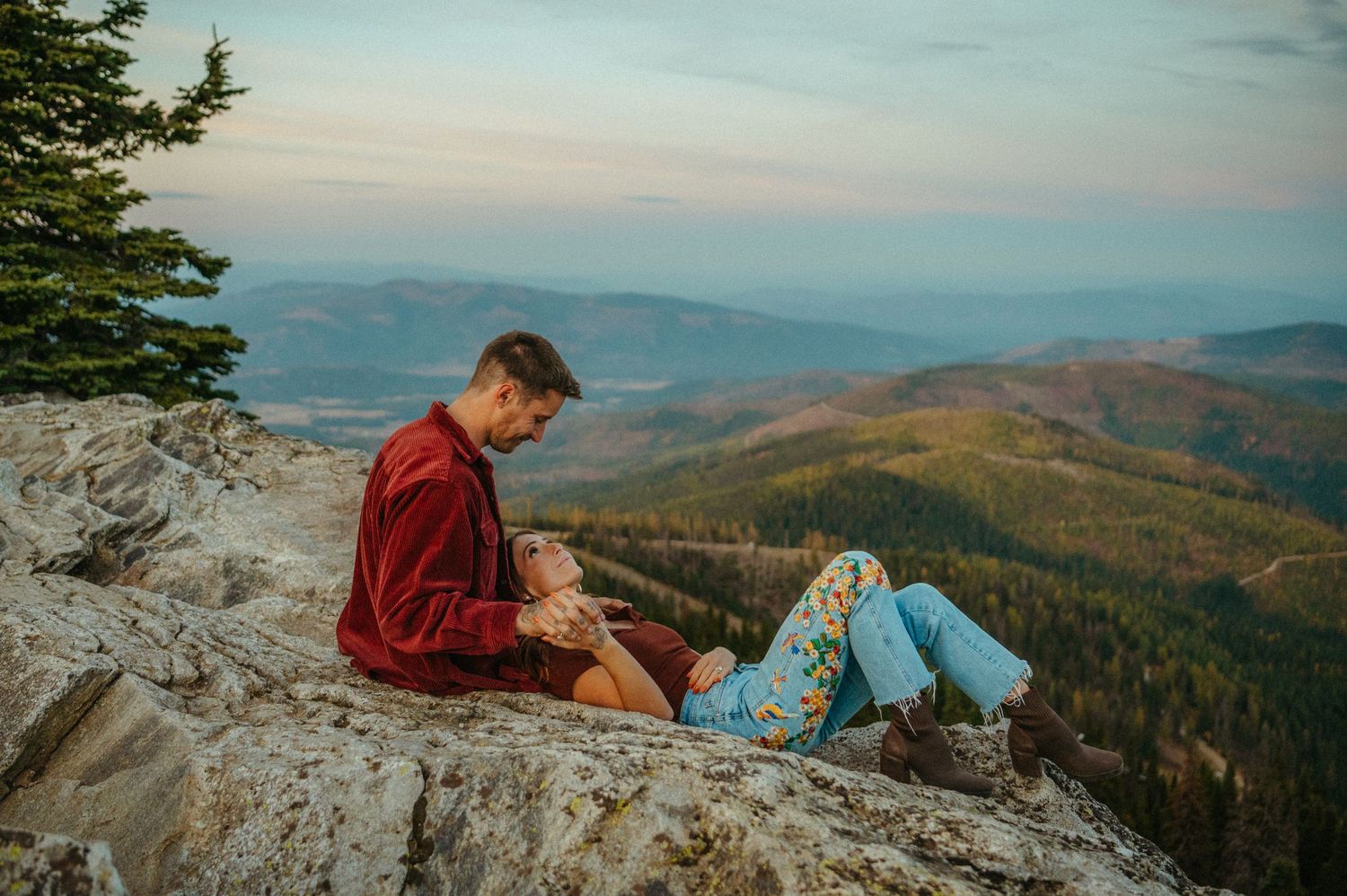 A couple relaxes together on a rocky mountain overlook with scenic forest valleys stretching into the distant horizon.