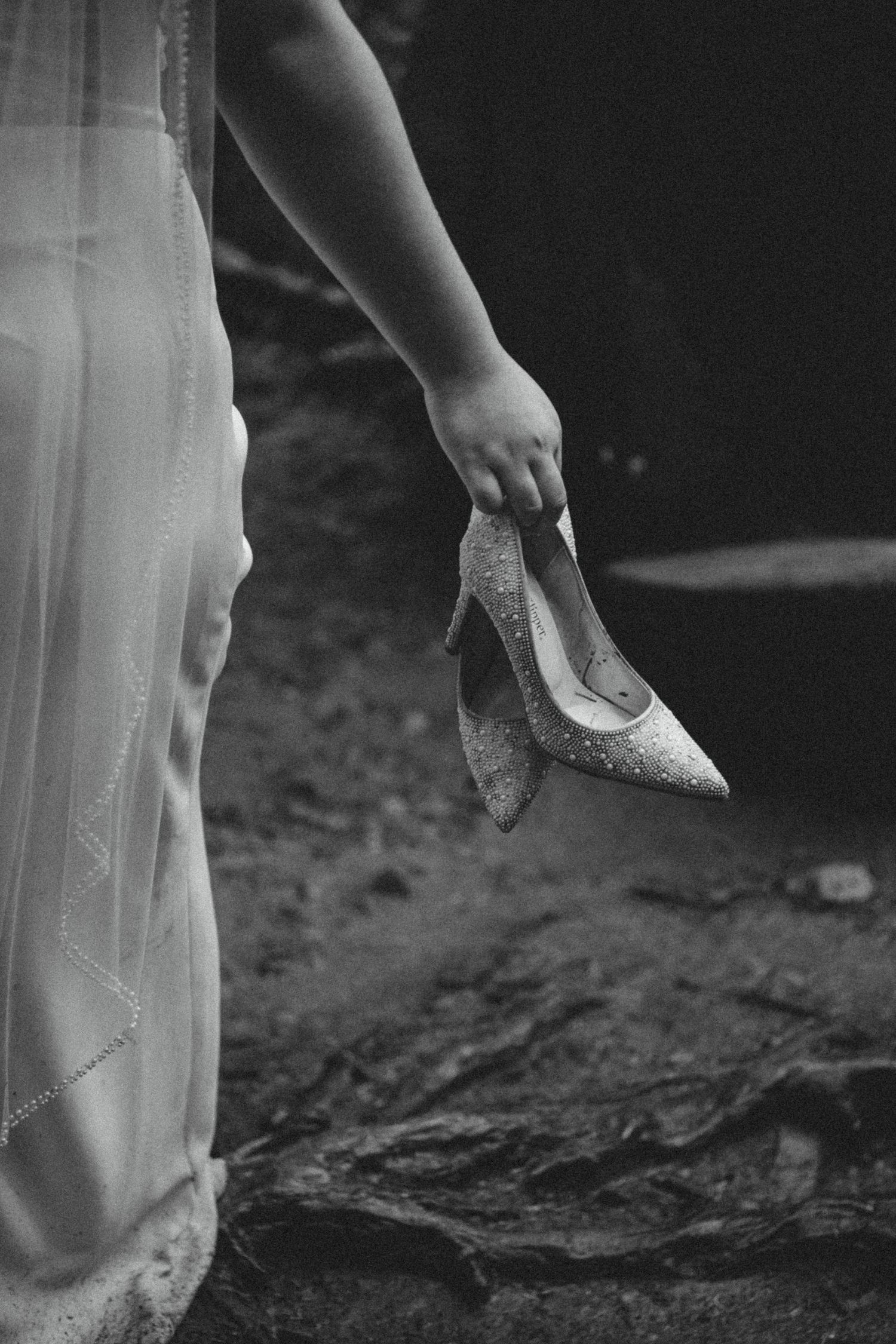 A hand holding a glittery high heel shoe against a flowing white dress in black and white photography.
