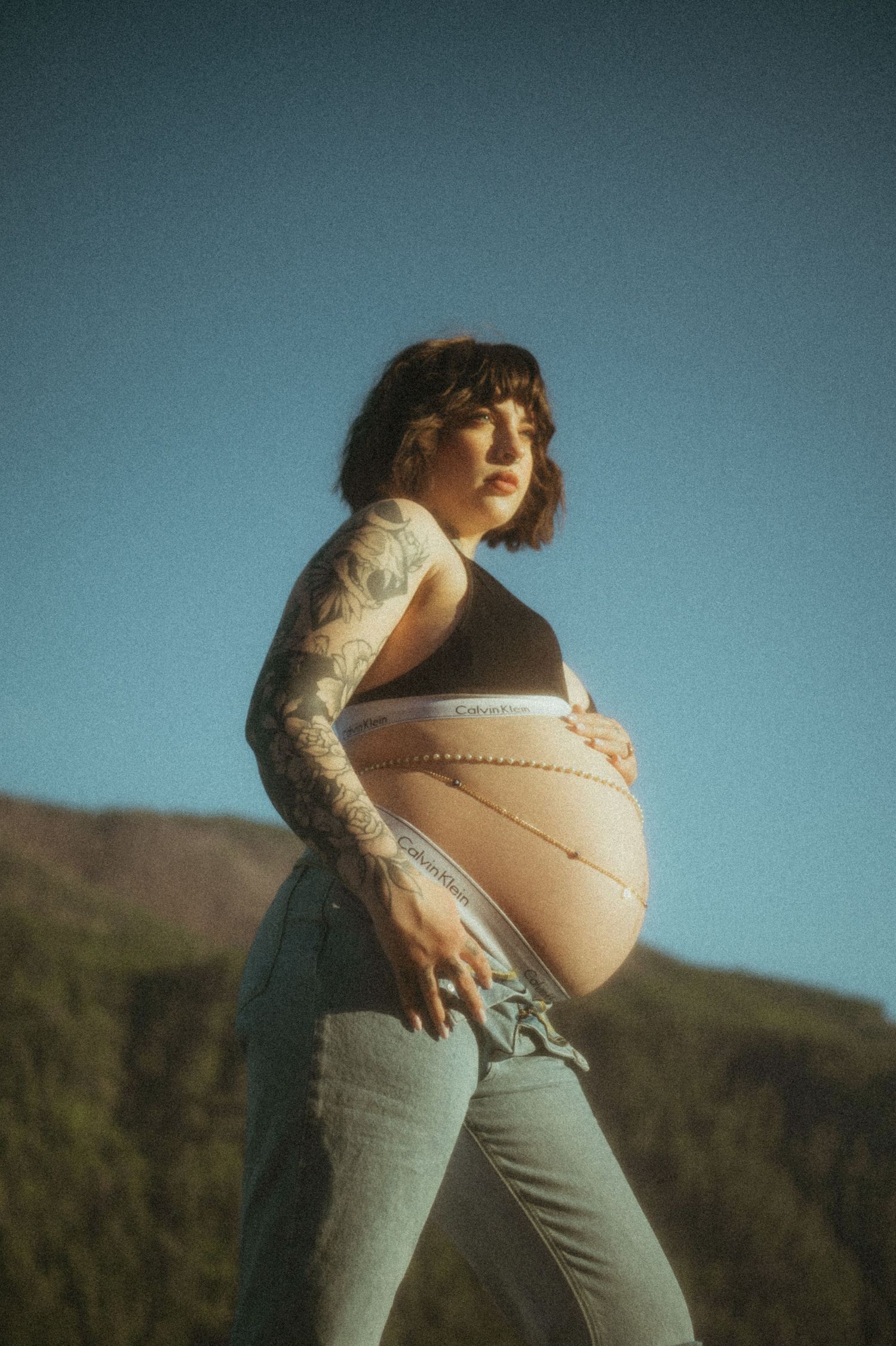 Art portrait in vintage style showing a person with tattoos wearing light wash denim and crop top against mountainous backdrop.