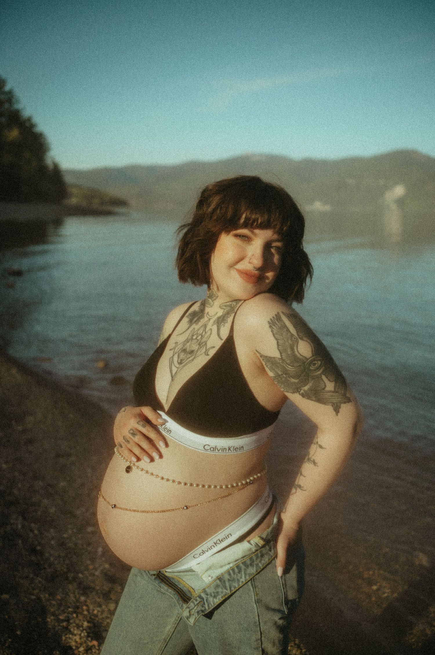 A tattooed pregnant person poses along a serene lakeshore with mountains in the background during sunset.
