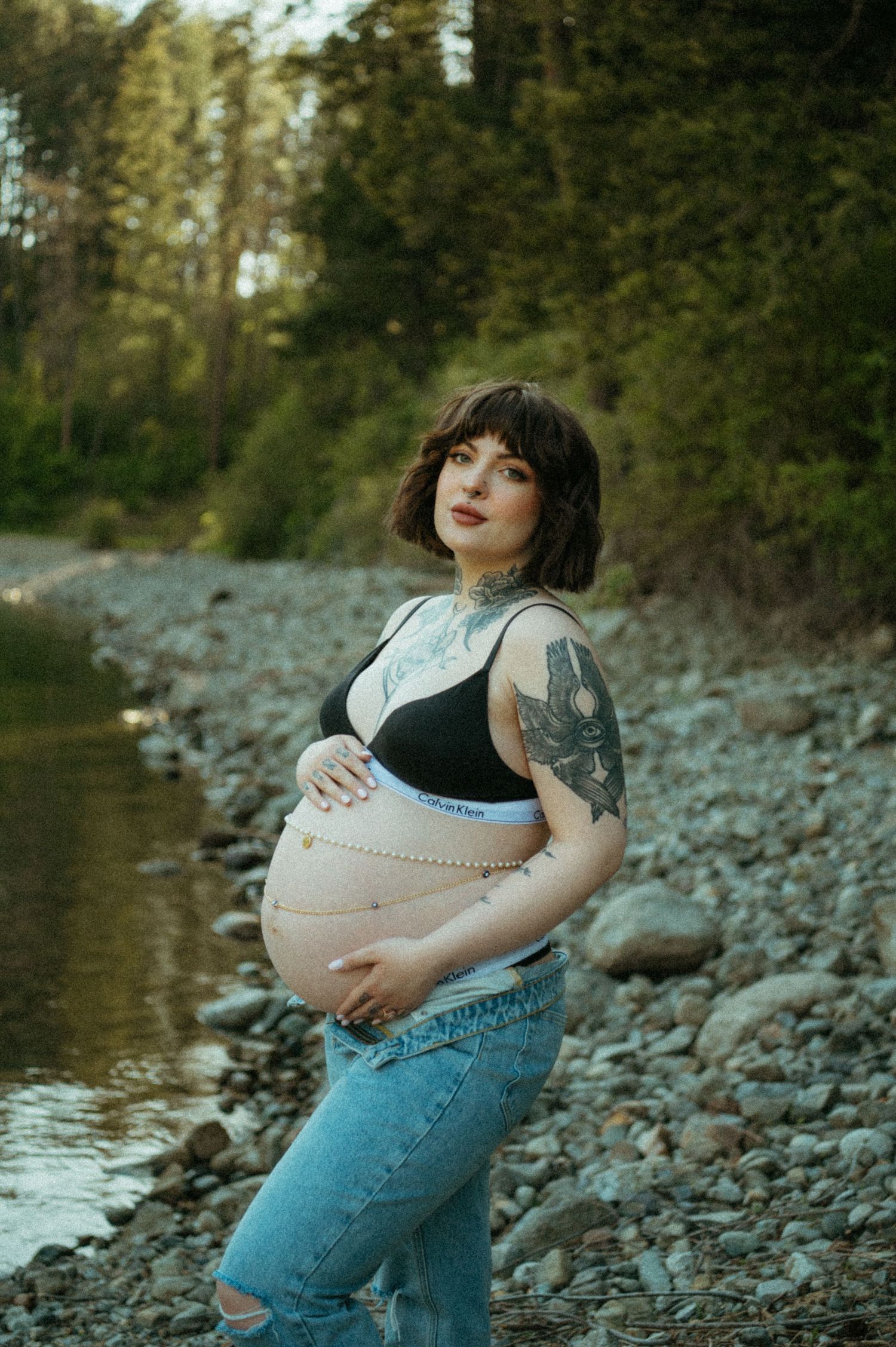 Expectant person in casual denim poses by a rocky riverbank during golden hour with trees in the background.