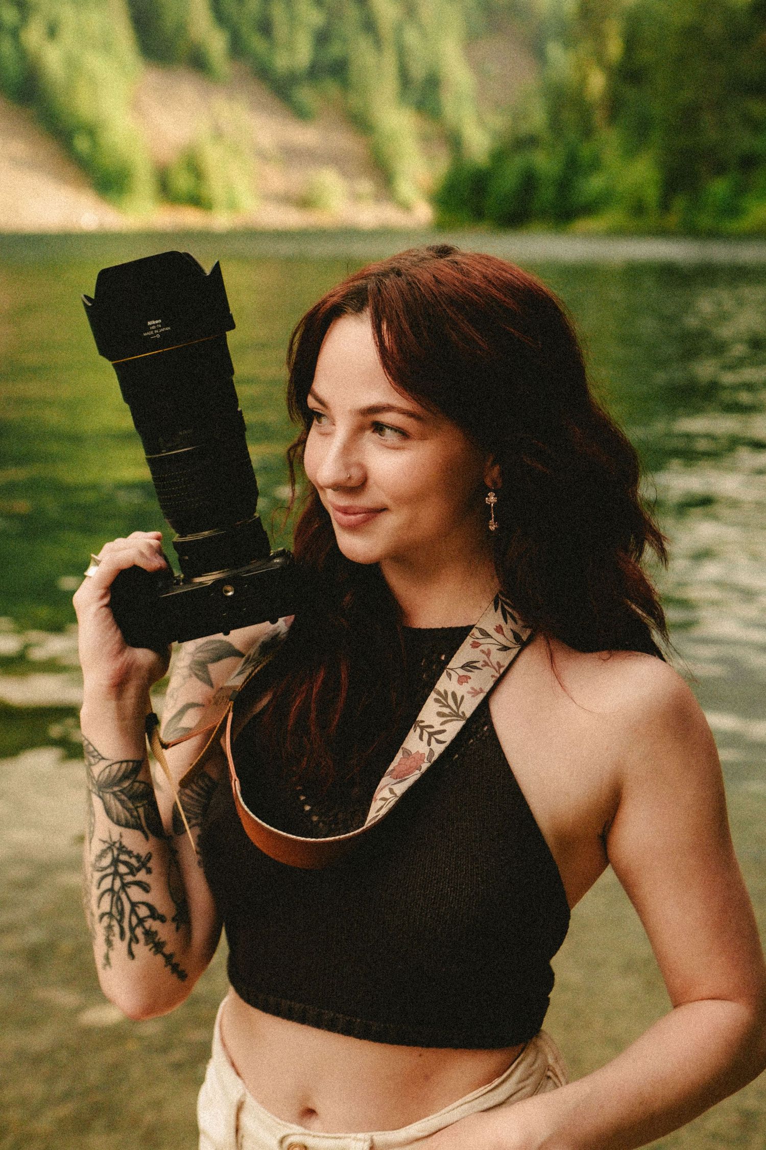 A photographer in a black crop top holding a professional camera near a lake with mountain scenery in the background.