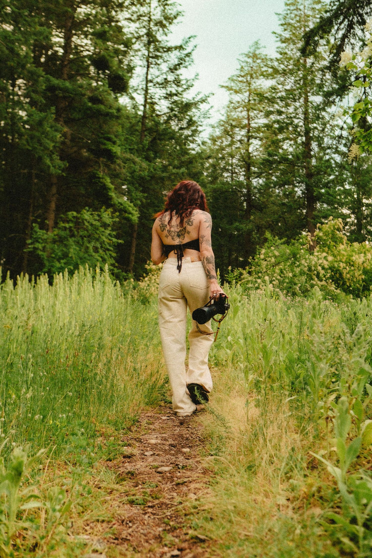 A photographer wearing beige pants walks along a forest trail carrying camera equipment through tall grass and evergreens.