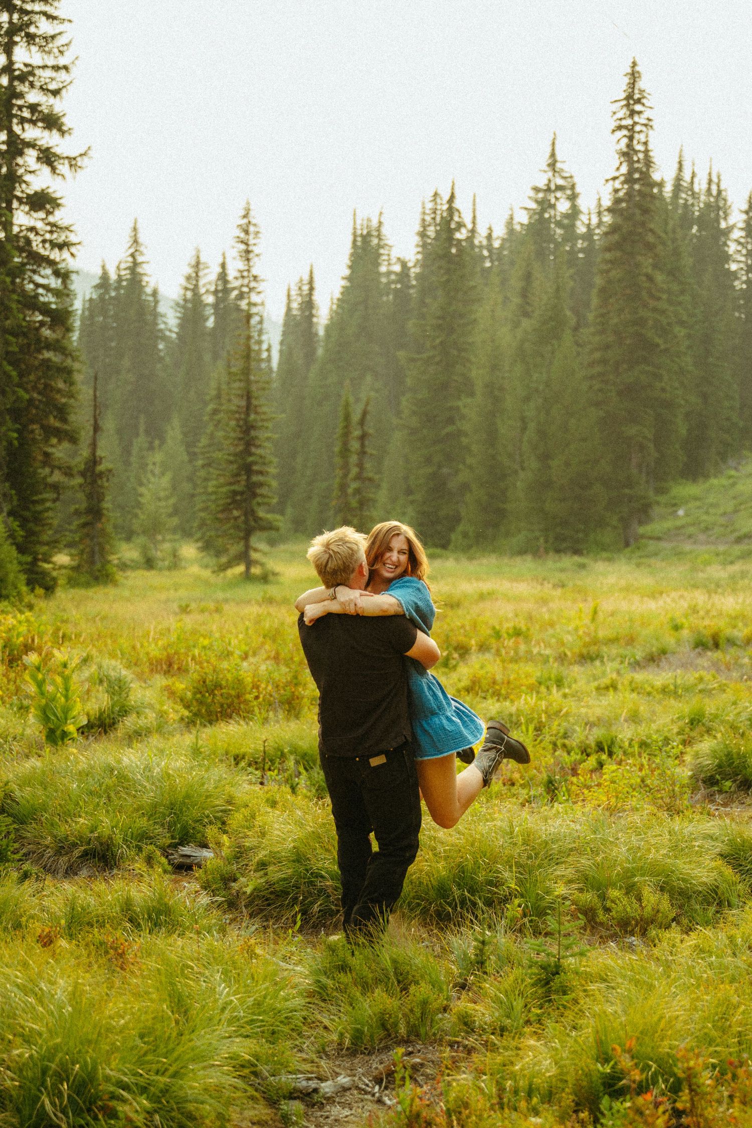 Two people embrace in a lush mountain meadow surrounded by tall evergreen trees in golden afternoon light.