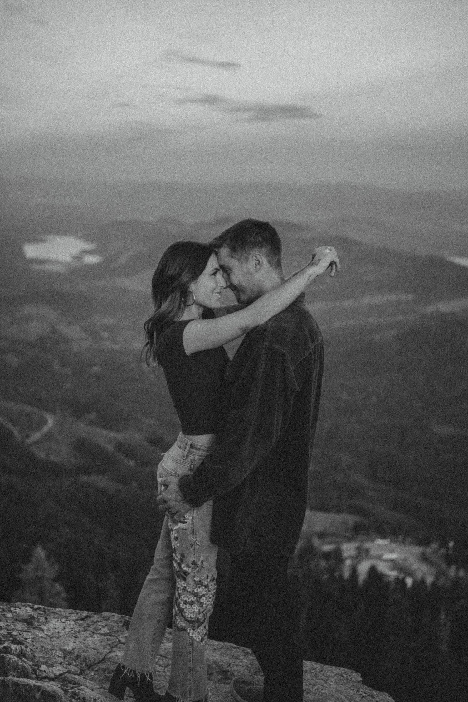 A romantic embrace captured in black and white on a mountaintop overlooking a vast mountainous landscape at dusk.