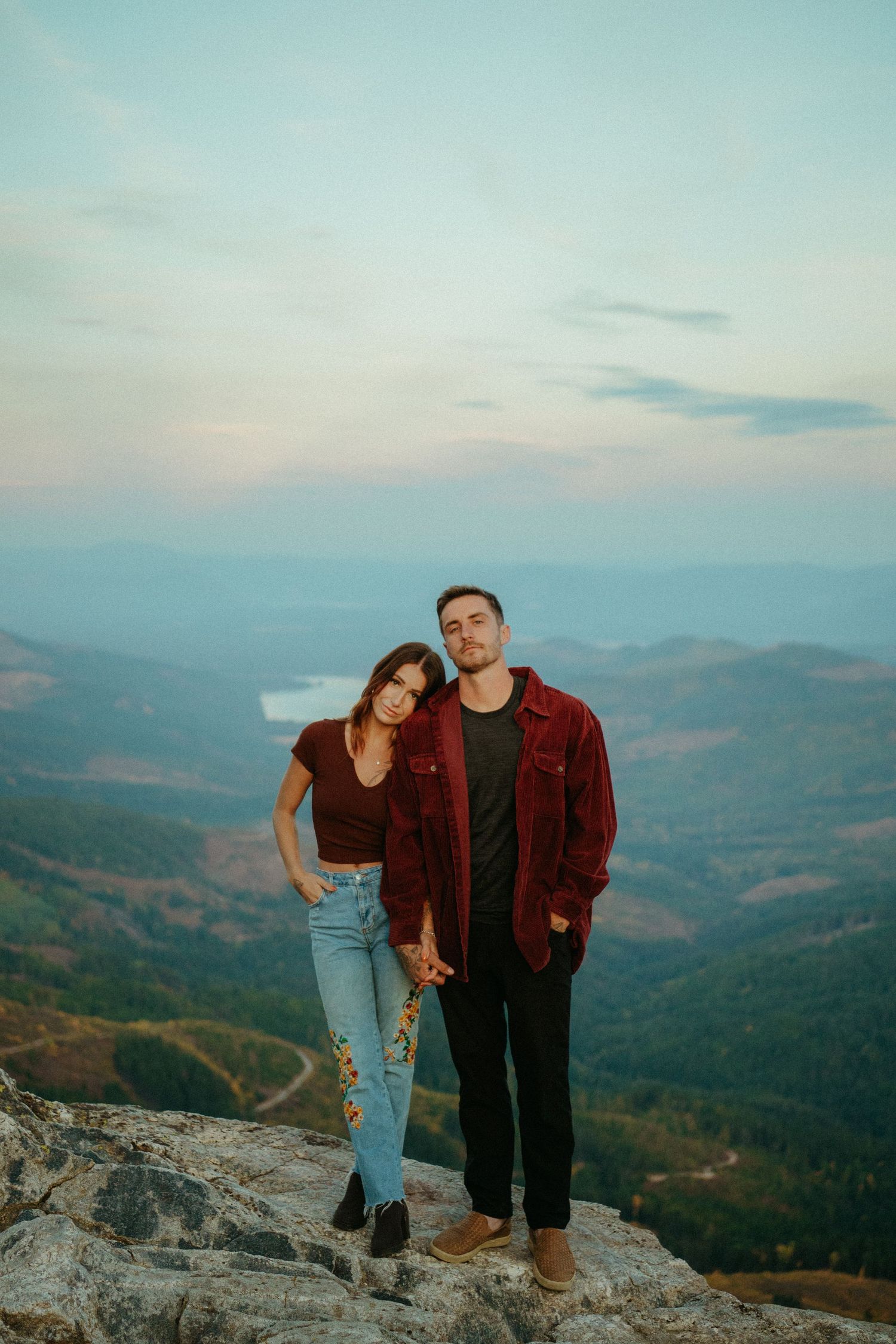 A couple stands together on a mountaintop overlook during sunset, with rolling hills and valleys stretching into the distance.