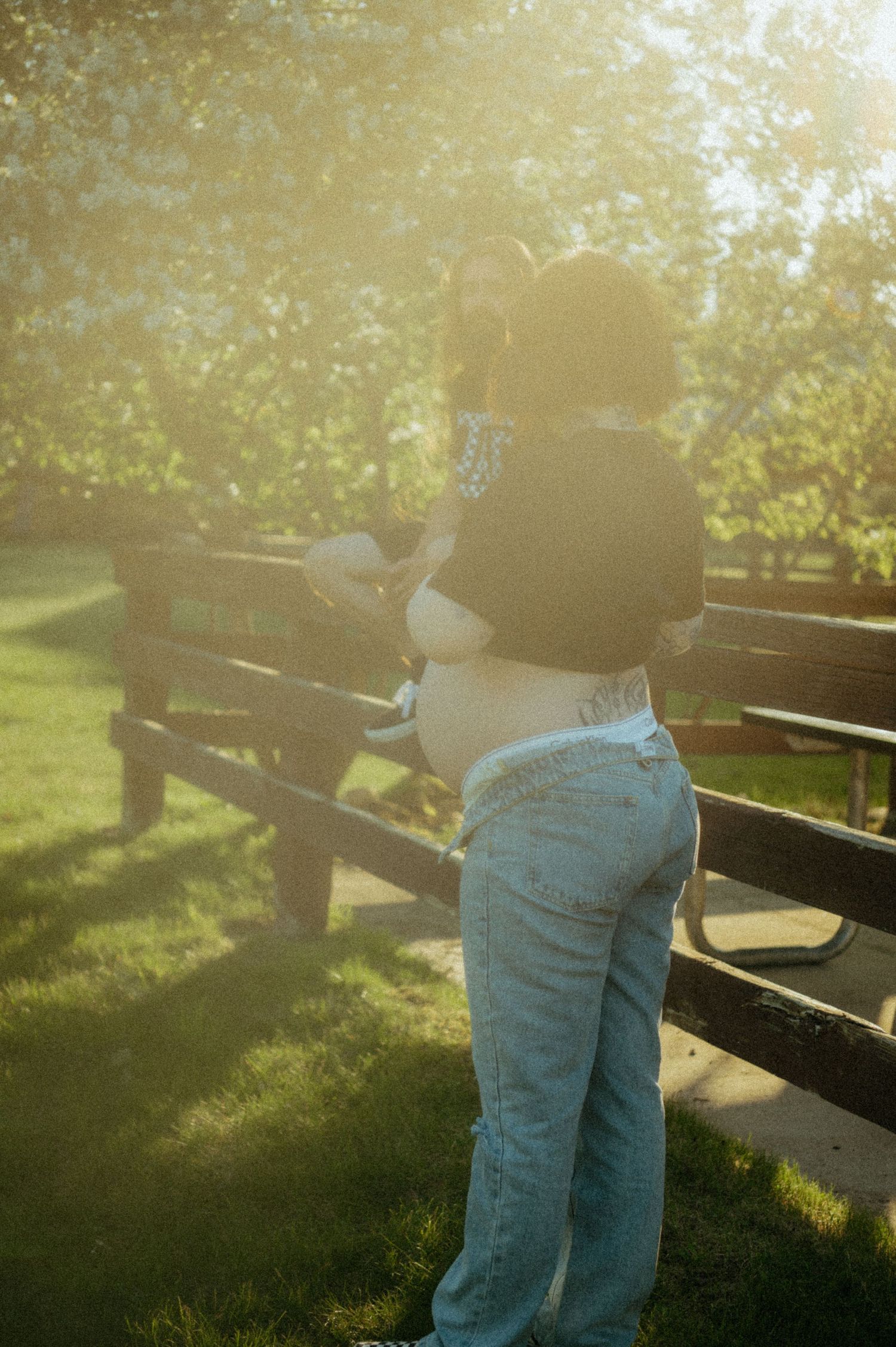 A person in casual attire leans against a wooden fence during golden hour with sunlight filtering through trees.