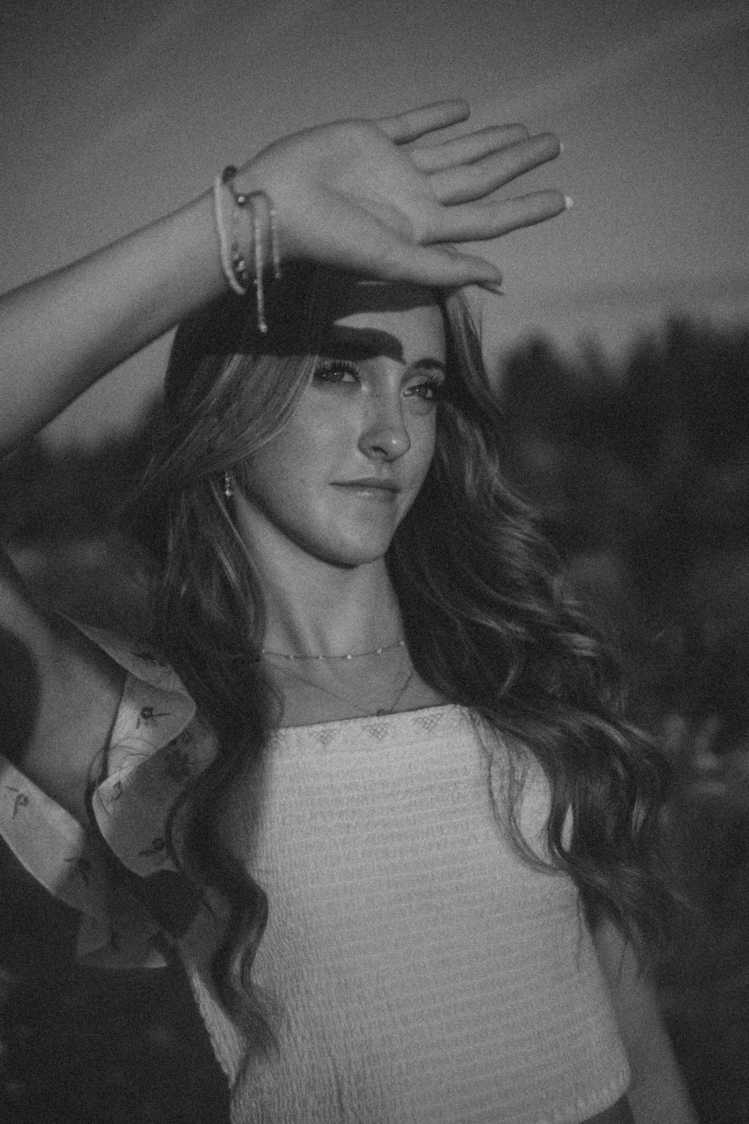 Artistic black and white portrait of woman in sleeveless top with hand raised to forehead in casual outdoor pose.