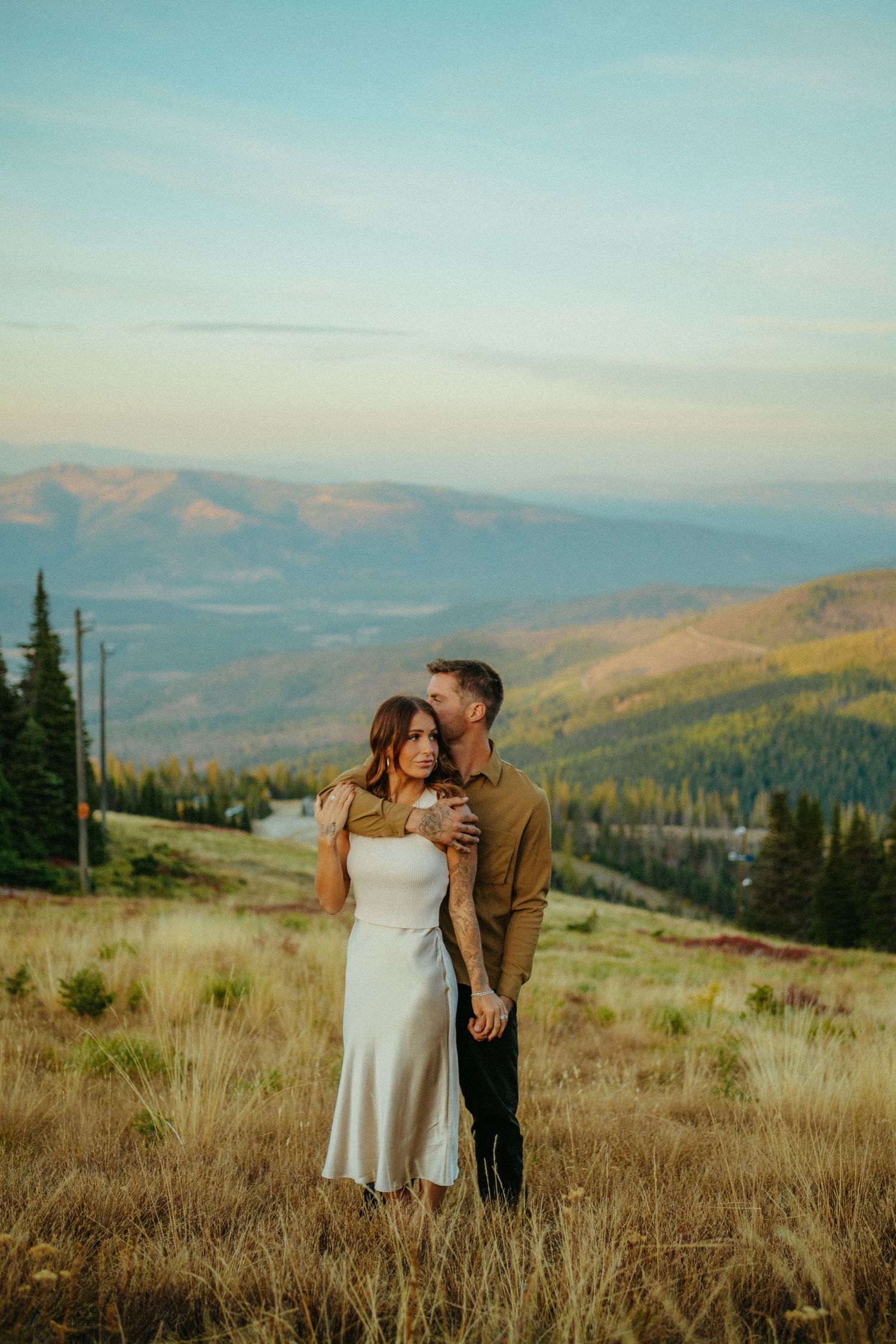 A couple embraces on a mountainside meadow with evergreen forests and rolling hills stretching into the distance at sunset.