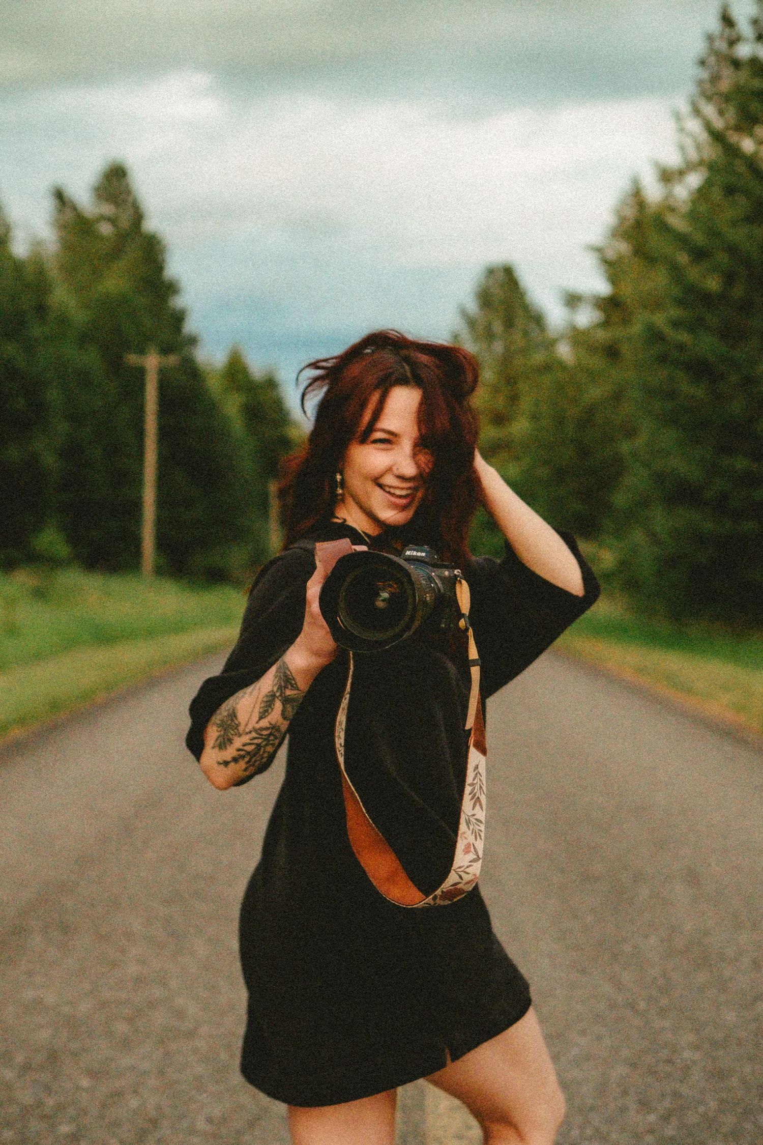 A photographer in a black dress holds a professional camera on a rural road surrounded by evergreen trees.